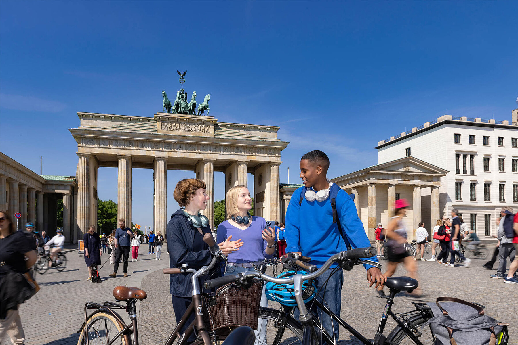 Drei Personen mit Fahrrädern unterhalten sich vor dem Brandenburger Tor. 