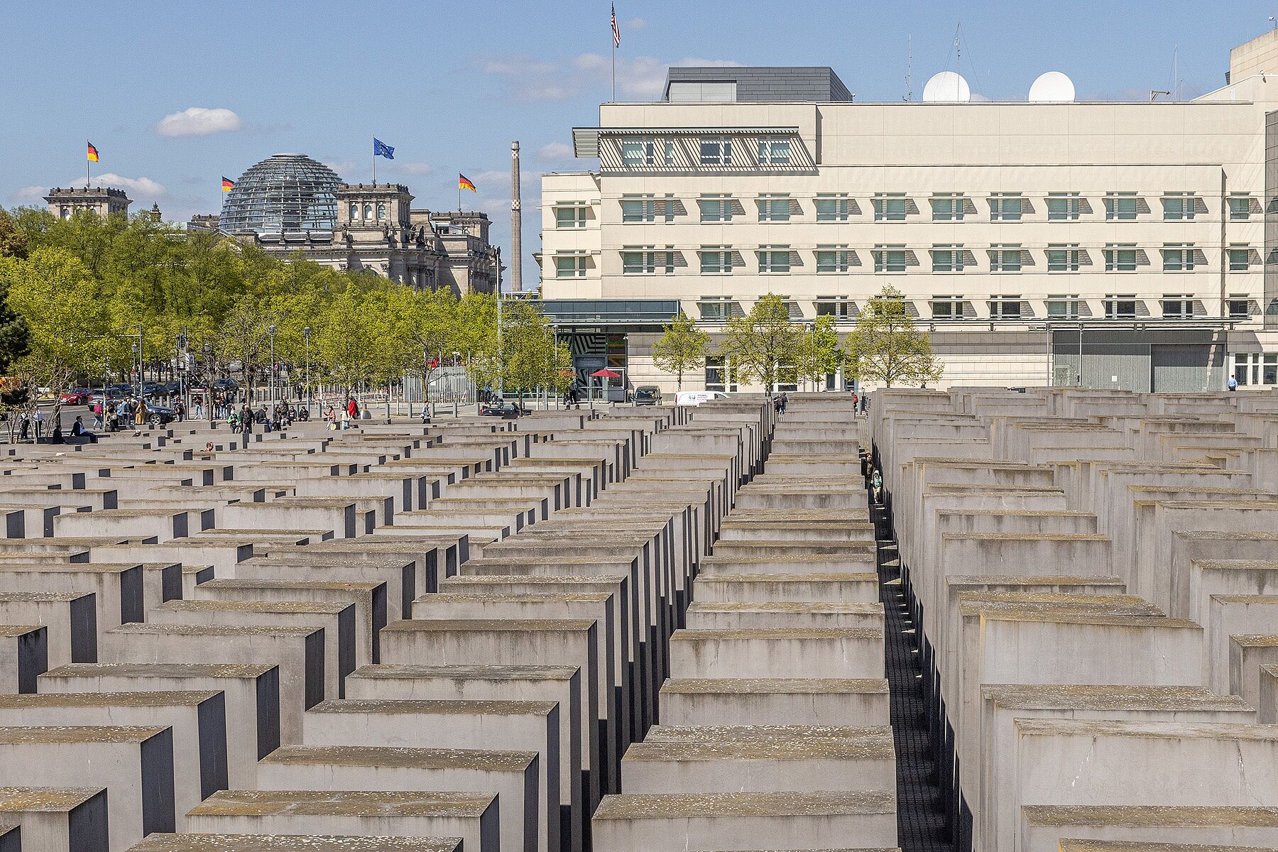 Im Vordergrund befinden sich die Betonstelen des Denkmals, links im Hintergrund das Reichstagsgebäude mit Kuppel, rechts hinten das weiße Gebäude der Botschaft der USA.