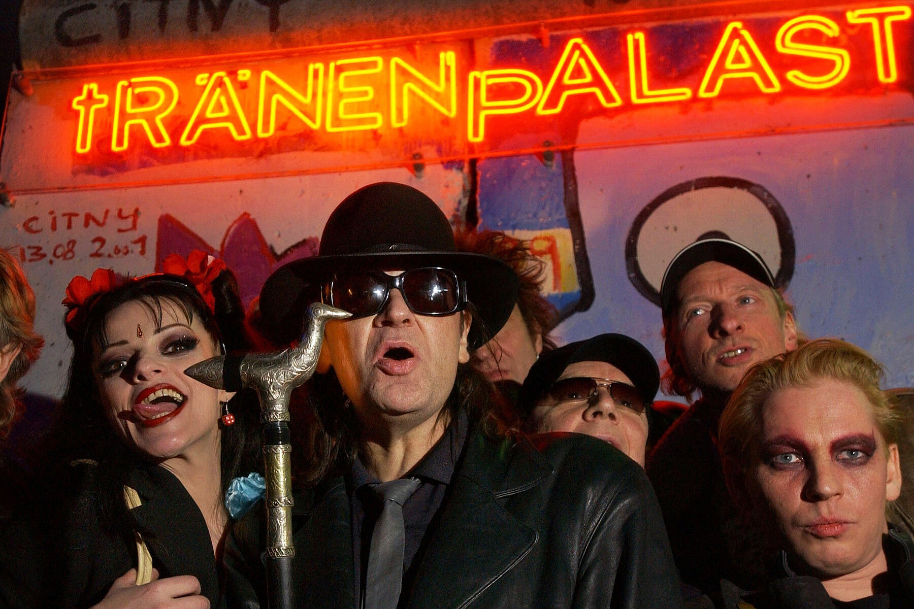 Nina Hagen, Udo Lindenberg, Ben Becker und weitere Prominente vor der roten Leuchtschrift tRÄNENpALAST auf einem Stück der Berliner Mauer mit Graffiti. 