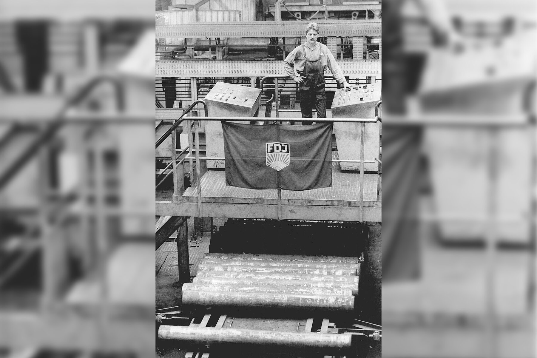 Arbeiter auf einem erhöhtem Metallbalkon. Am Geländer vor ihm hängt eine Fahne mit dem Logo der Freien Deutschen Jugend. Davor und darunter befinden sich mehrere Metallrohre in einer Reihe.