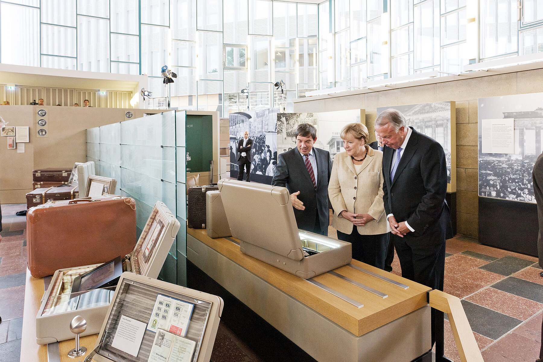 Koffer mit Museumsobjekten auf Erhöhungen in der Ausstellung. Rechts Hans Walter Hütter, Angela Merkel und Bernd Neumann. 