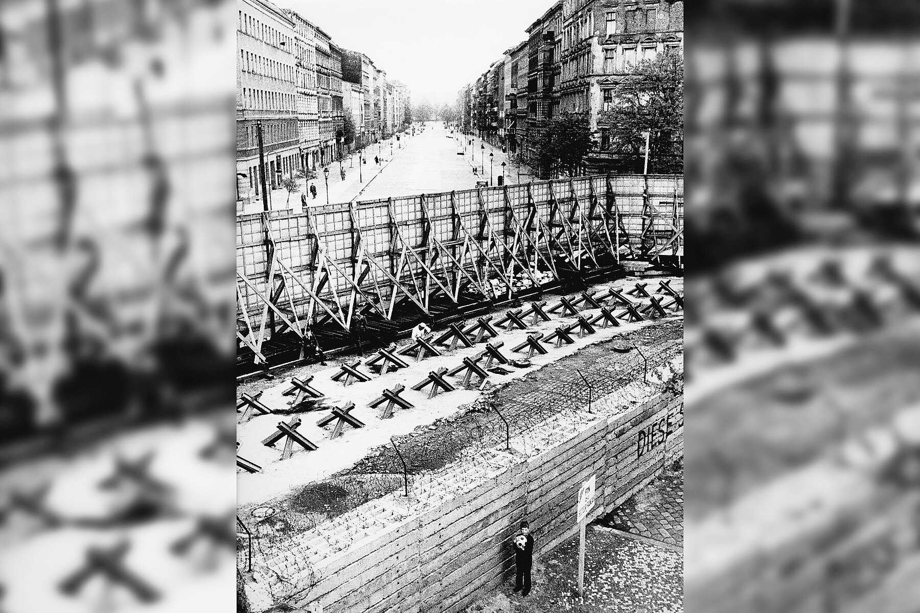 Ein kleiner Junge vor der Mauer. Im Hintergrund eine durch die Grenzanlage durchschnittene Straße.