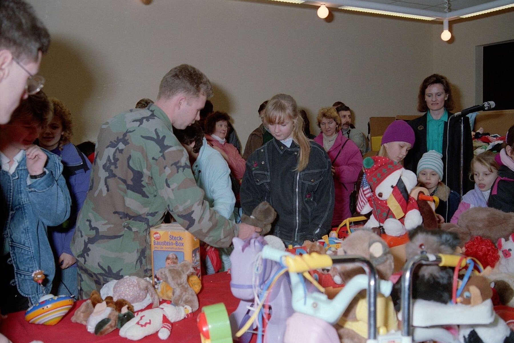 Ein amerikanischer Soldat umgeben von Geflüchteten aus der DDR. Im Vordergrund steht ein Tisch mit verschiedenen Spielzeugen.