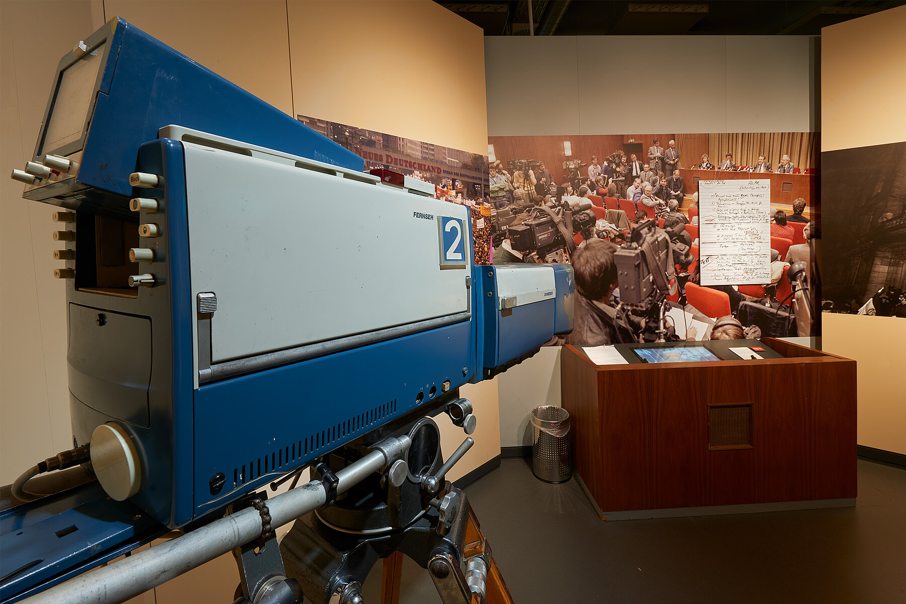 Eine Fernsehkamera links im Bild ist auf das Pult der Schabowski-Pressekonferenz gerichtet. Dahinter ein großformatiges Bild der Konferenz und ein vergrößerter Druck von Schabowskis Notizzettels von der Pressekonferenz an der Wand. 