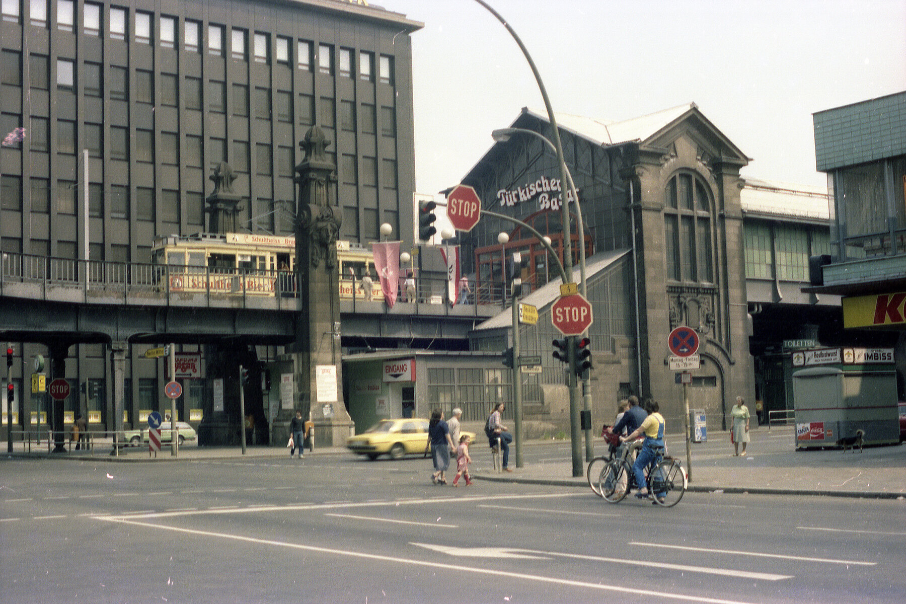 Der Hochbahnhof Bülowstraße mit Schriftzug Türkischer Basar, Gleisen und historischer Straßenbahn hinter einer Straßenkreuzung mit Fahradverkehr, Fußverkehr und Autos. 