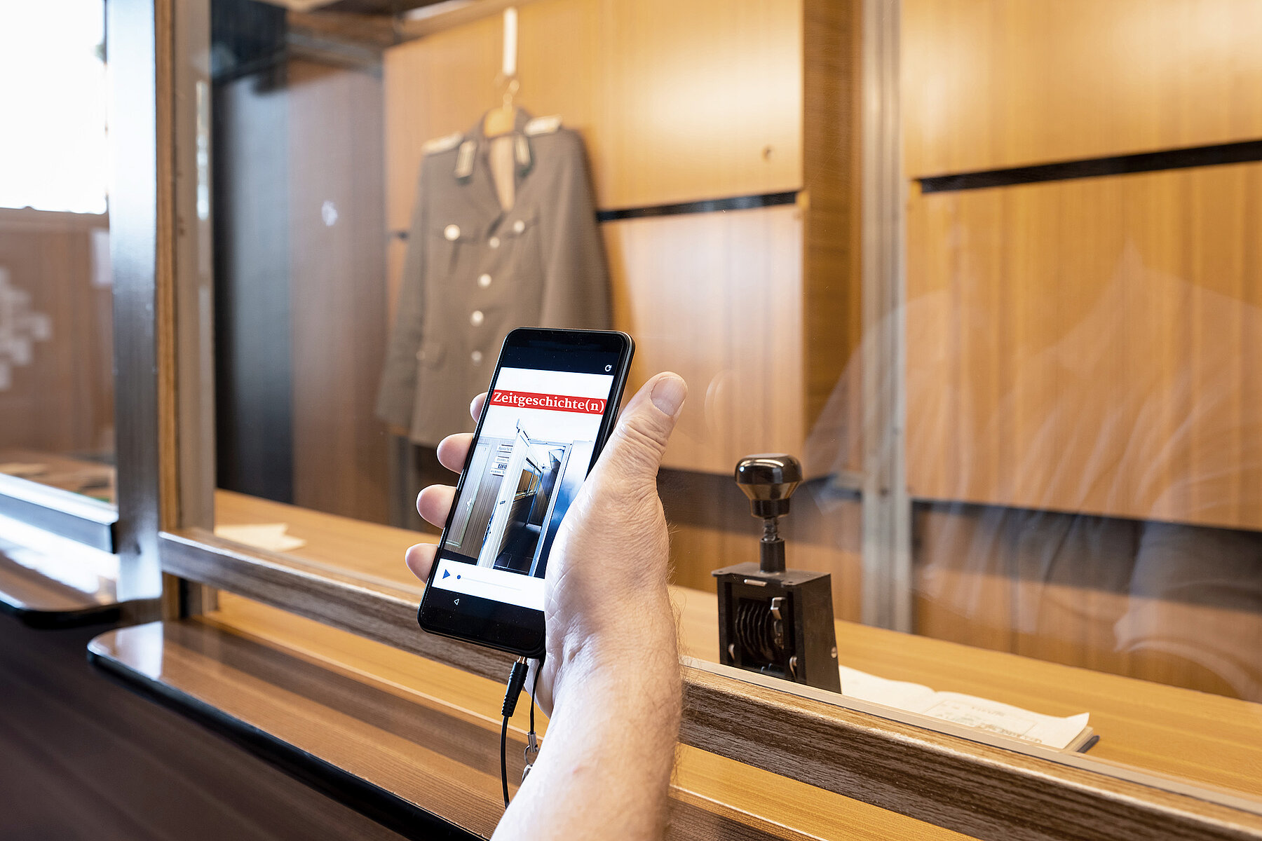 Grenzkontrollkabine in der Ausstellung im Tränenpalast. Eine Hand hält ein Smartphone, das den Museumspodcast zeigt. Hinten, durch eine Glasscheibe getrennt, steht ein Stempel und hängt eine Uniform.