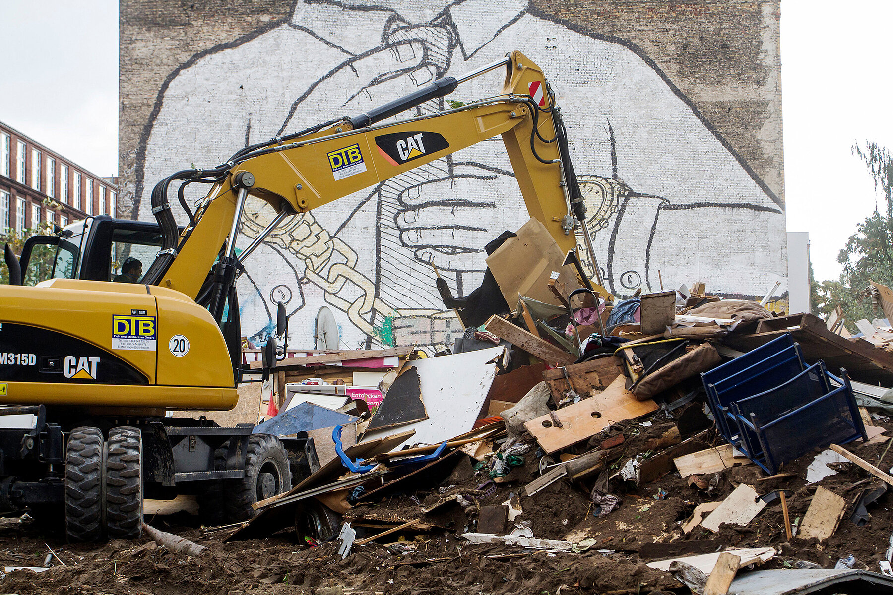 A yellow excavator, pushes bulky objects together on the Cuvry wasteland, behind it is a house wall with a mural of a person painted in white, wearing gold watches on both wrists, linked by a chain.
