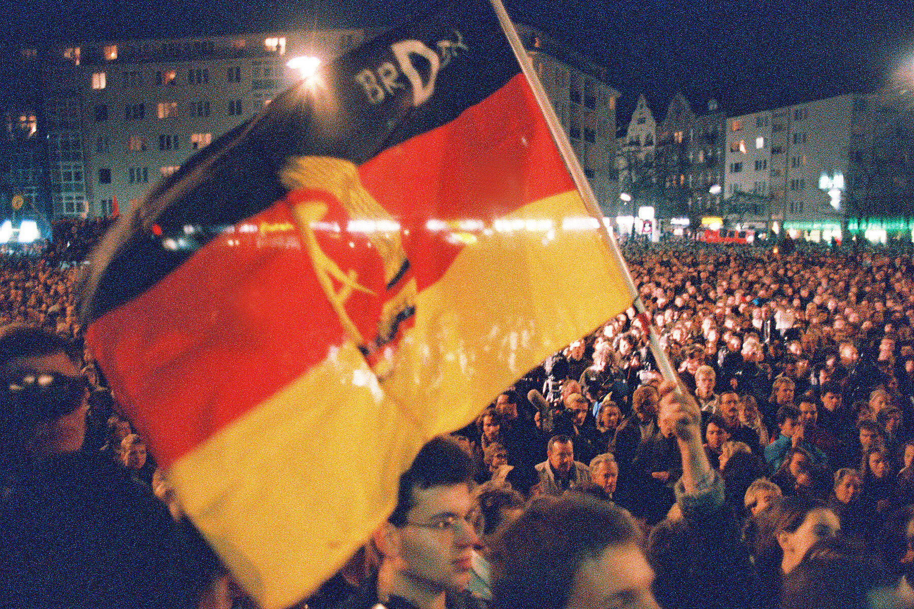 A huge crowd of people in front of Rathaus Schöneberg. In the foreground a demonstrator waves a flag with half of the GDR's national emblem and the inscription FRGDR.