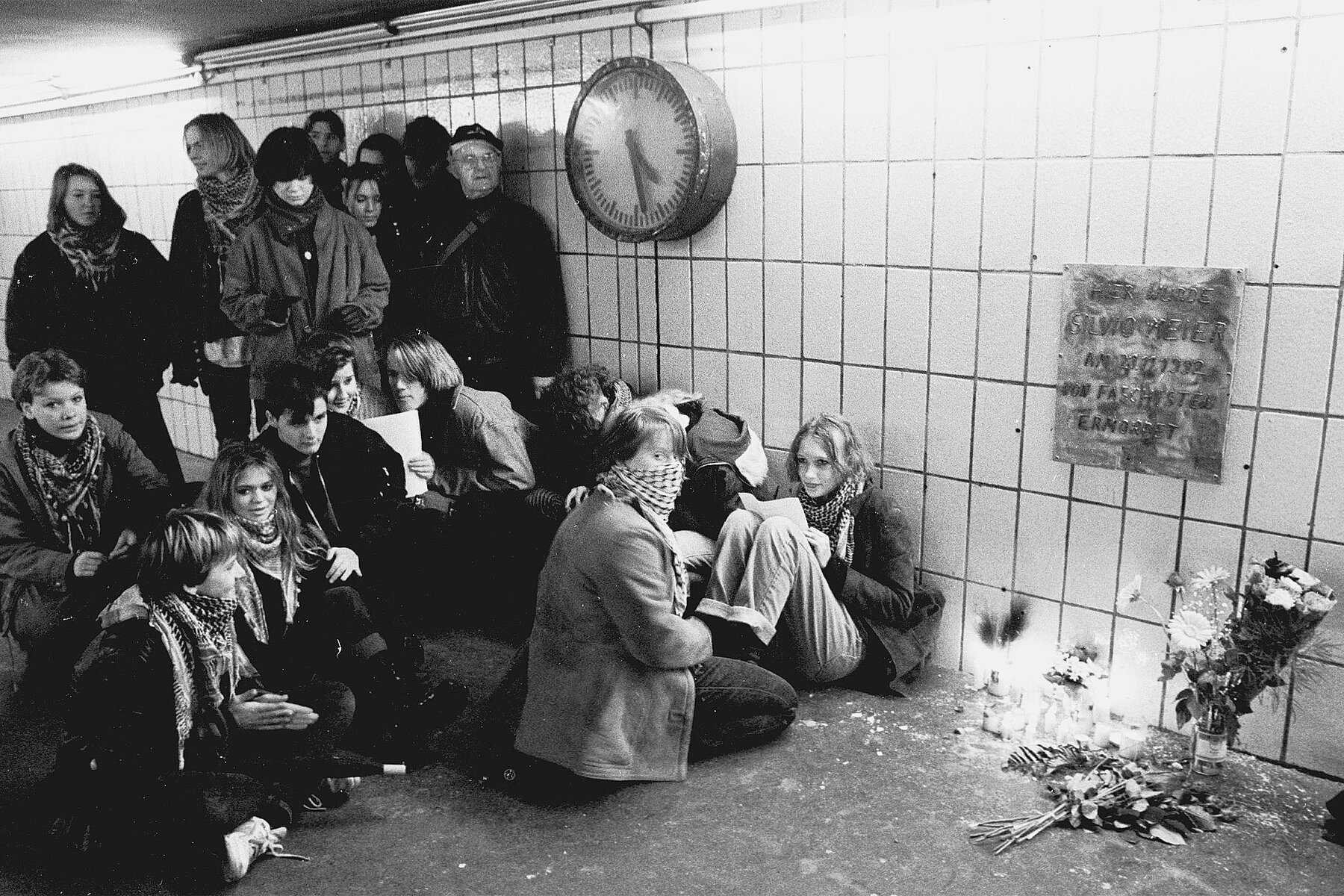 People stand and sit in front of the memorial plaque for Silvio Meier in the subway station. In front of the memorial plaque are flowers and candles.