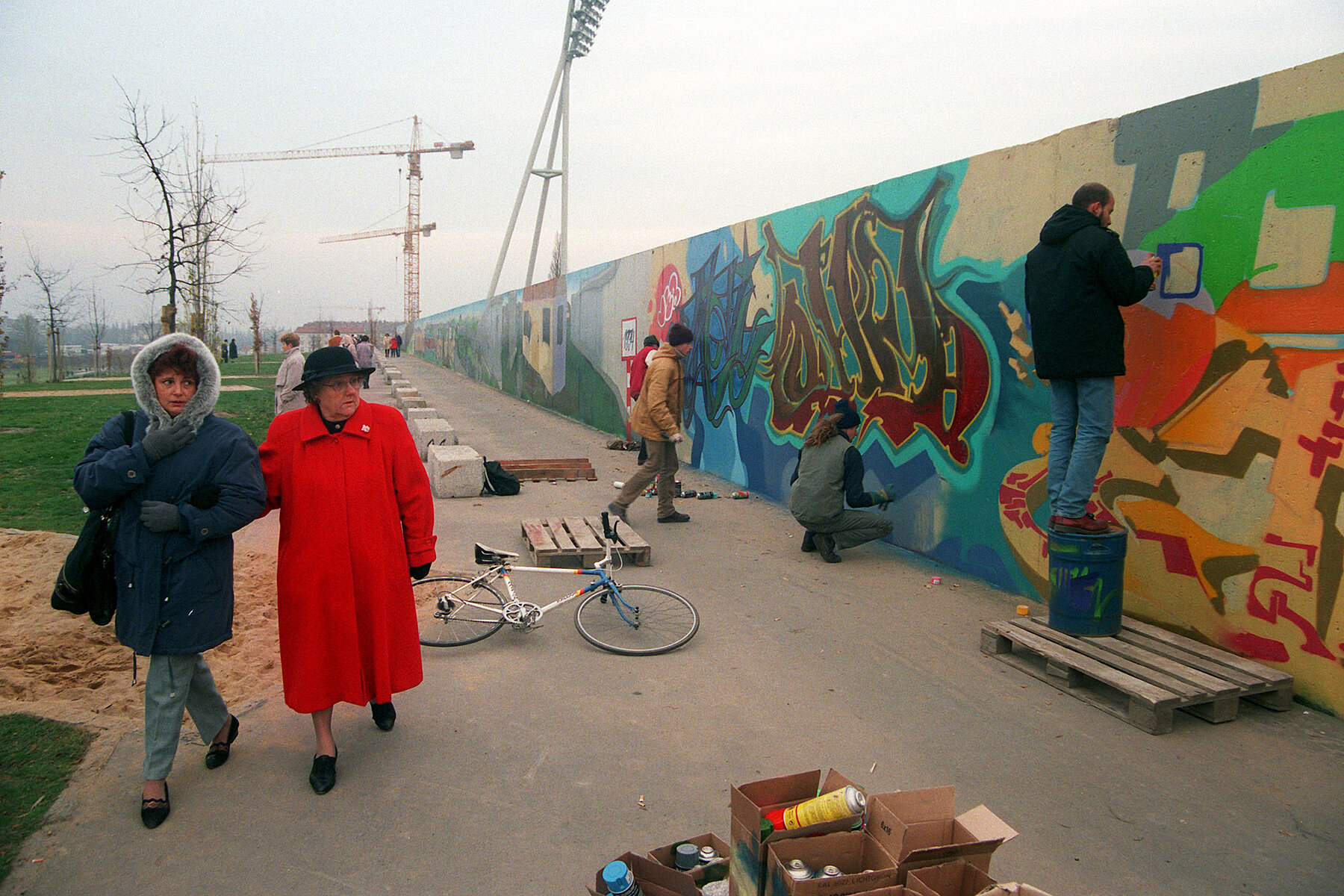 Sprayers paint the Wall between Mauerpark and Jahnsportpark, while two women walk past them. Construction cranes can be seen in the background.
