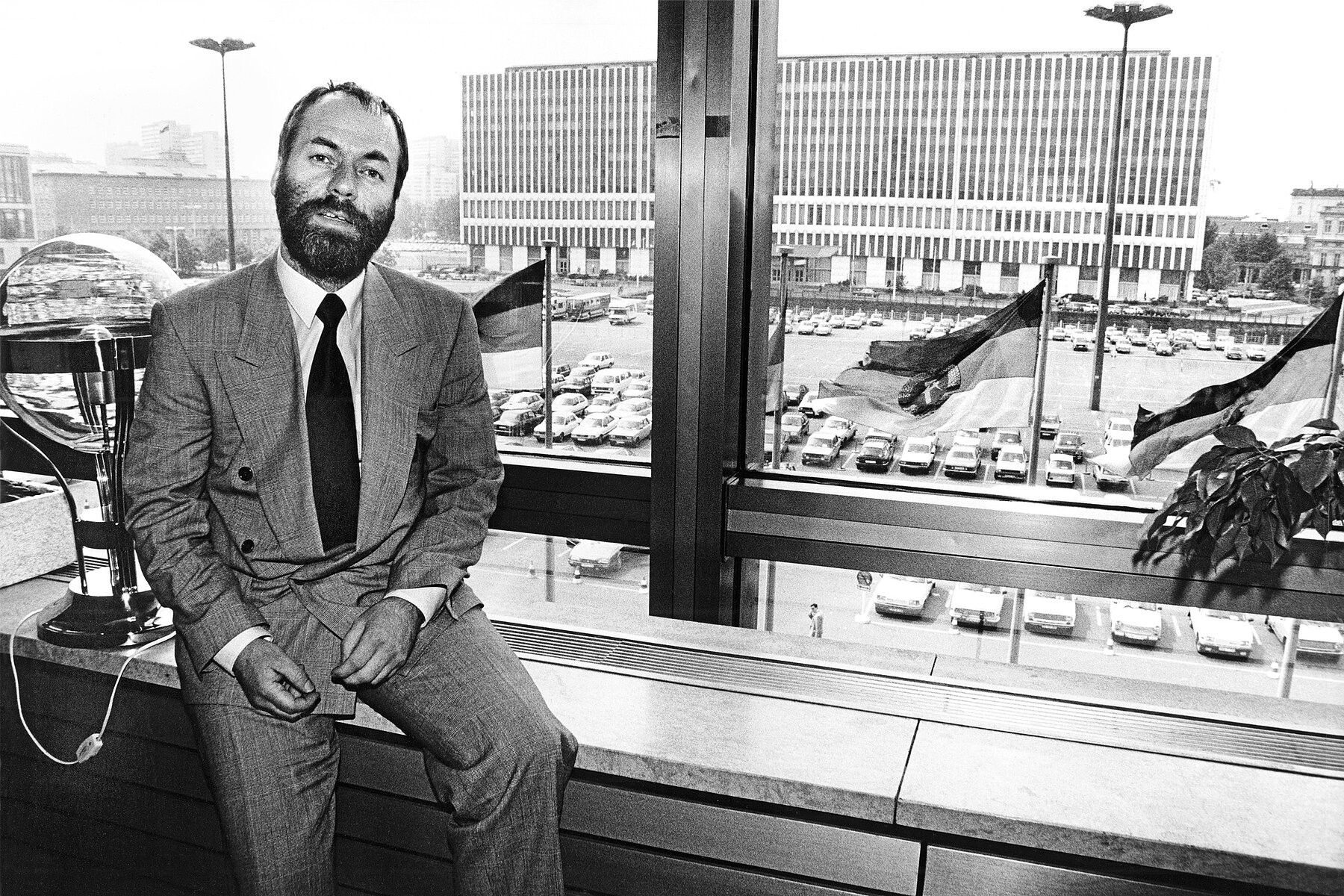 Markus Meckel sits on a windowsill in the Palace of the Republic. Behind him is a parking lot. Three GDR flags wave in the wind. Behind them is the white high-rise building of the Ministry of Foreign Affairs of the GDR.