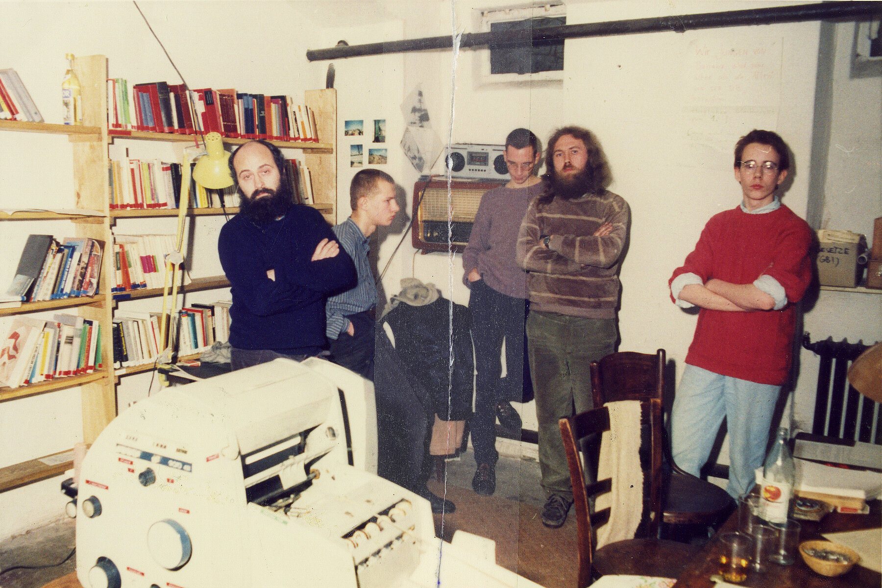 Five men ages 14 to 34 inside the Umweltbibliothek, with a book shelf behind them to the left and a printing machine in the foreground. 