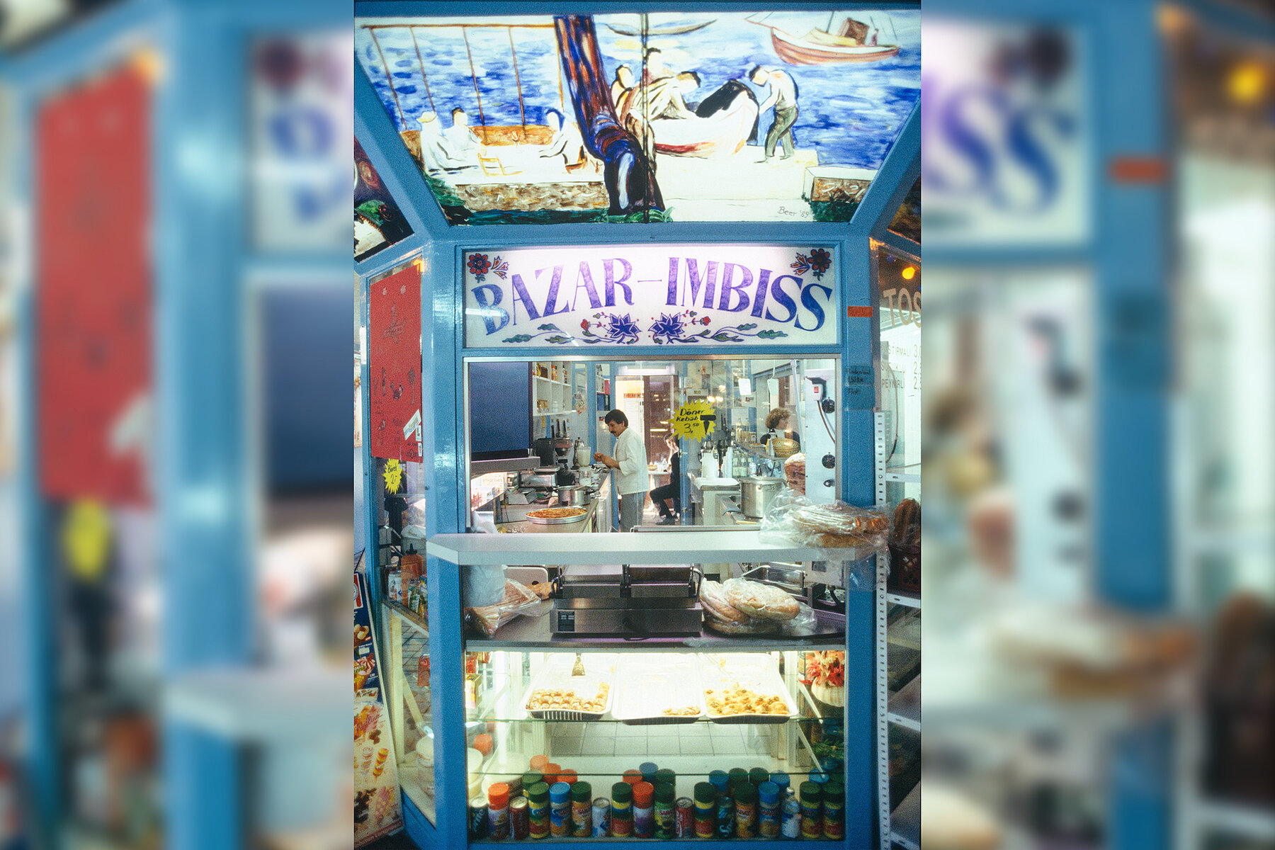 The counter of the Bazaar snack bar with pita bread and baklava in blue, above it a painting. Inside, people are working in the kitchen. 