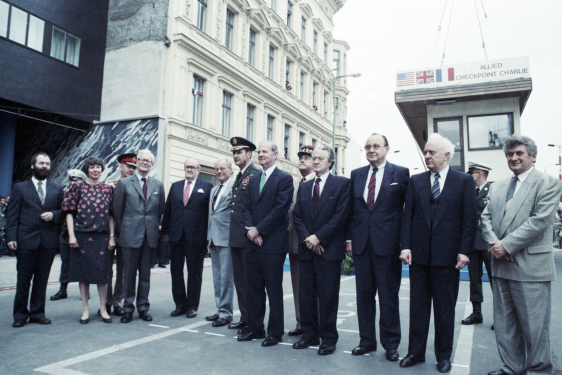 Männer in Anzügen und militärischen Uniformen und eine Frau stehen in einer Reihe. Hinter Ihnen hängt das Kontrollhäuschen an Seilen in der Luft.