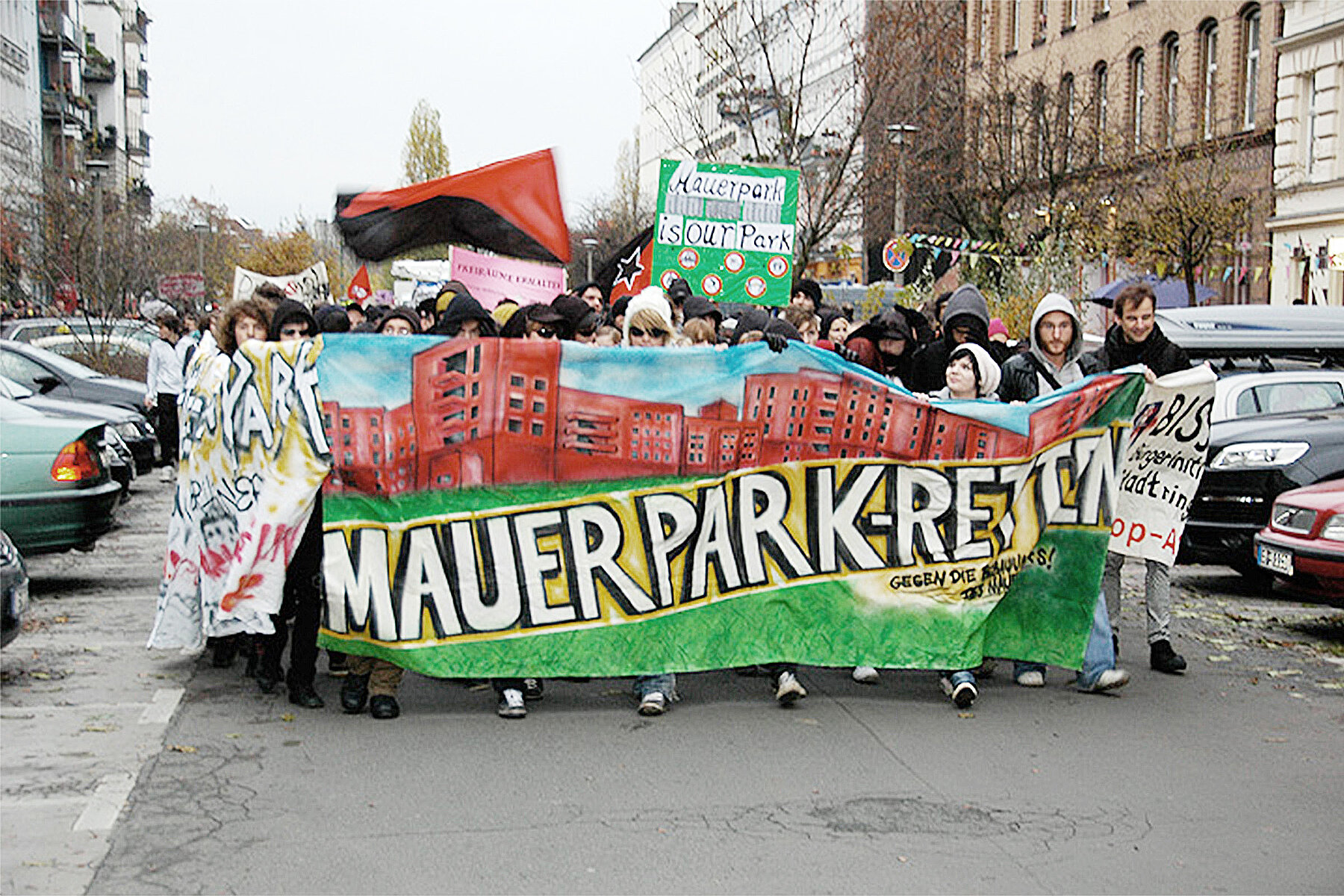 Demonstrierende laufen mit einem bunten Banner, auf dem Mauerpark retten steht, eine Straße entlang.