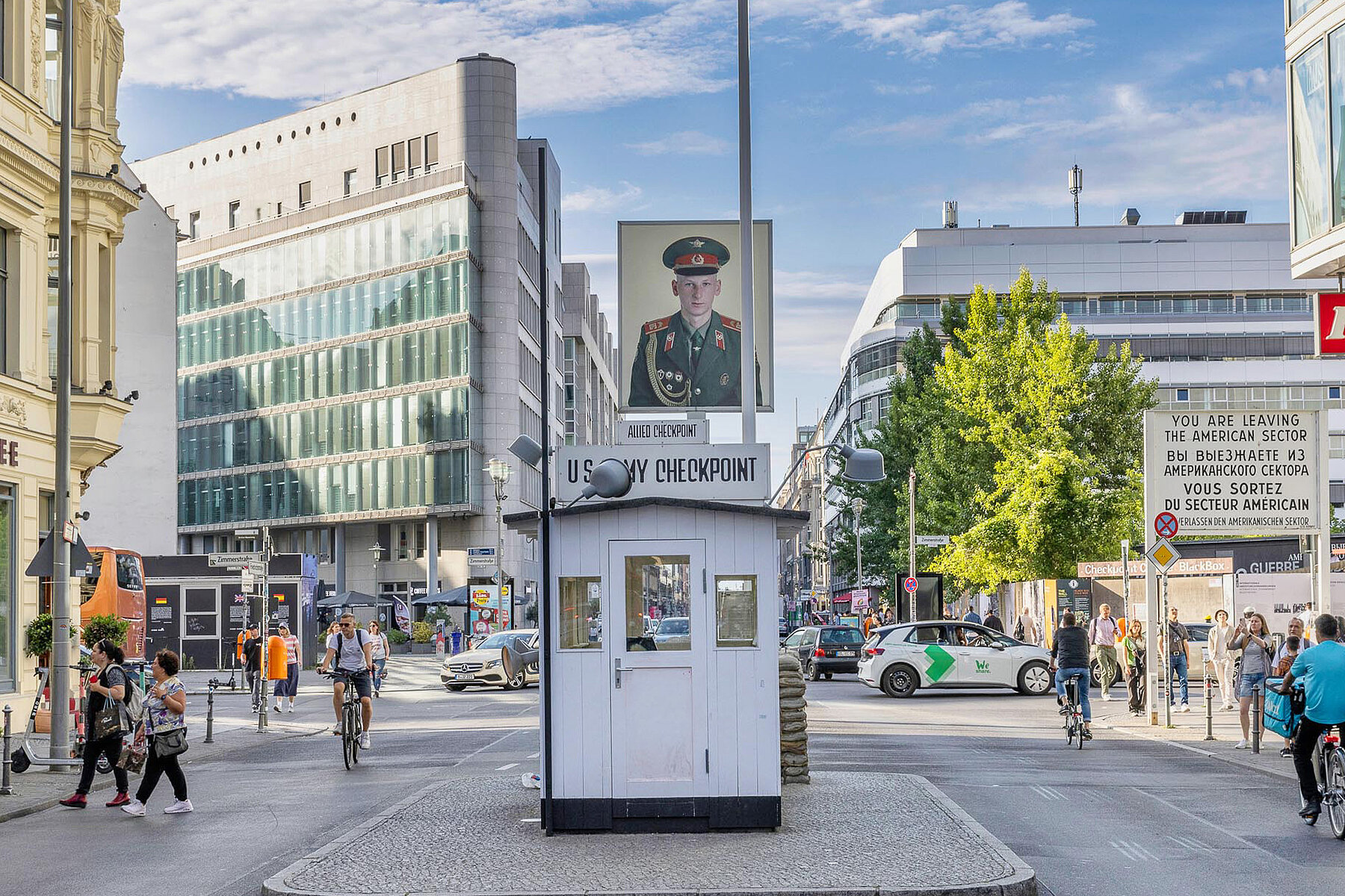 The control booth at Checkpoint Charlie stands in the middle of the street. People walk and ride by on bicycles. There are several new buildings on the side of the road.