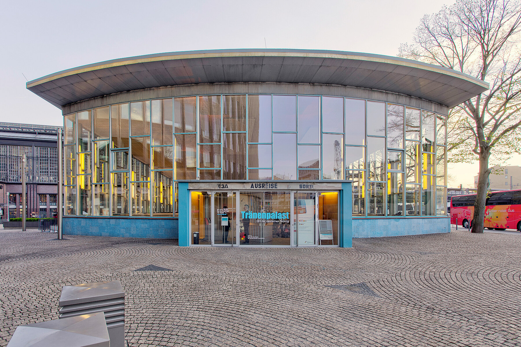 Der Tränenpalast in Frontalansicht mit Glasfront und Eingangstür vor dem Bahnhof Friedrichstraße im Dämmerlicht.