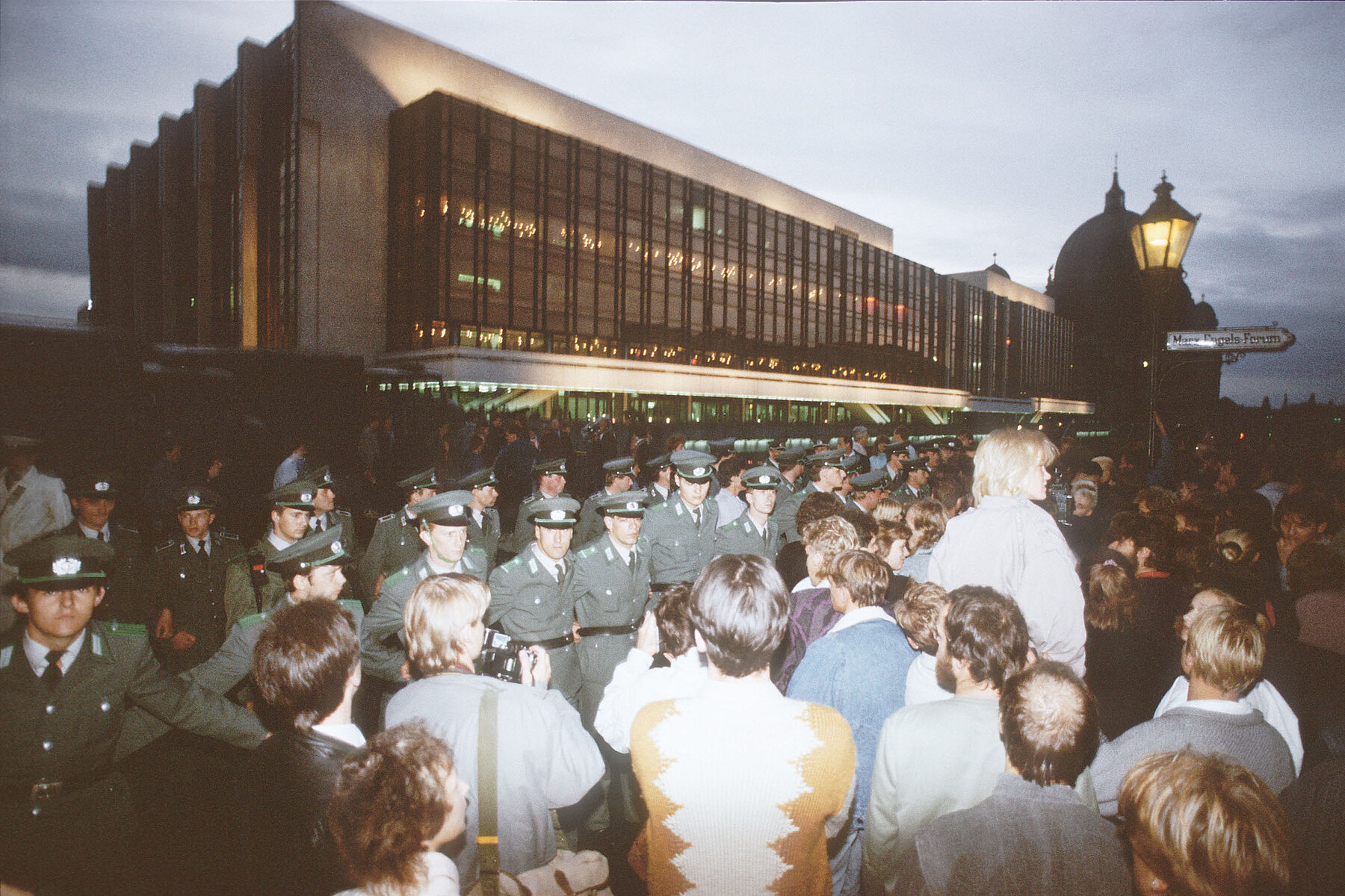 A group of people stands in front of the Palace of the Republic in the evening. Between them and the building are several rows of uniformed police.