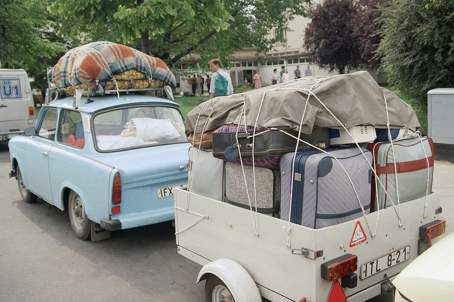 Ein hellblauer Trabi mit Dachgepäck und mit Koffern beladenem Anhänger. 