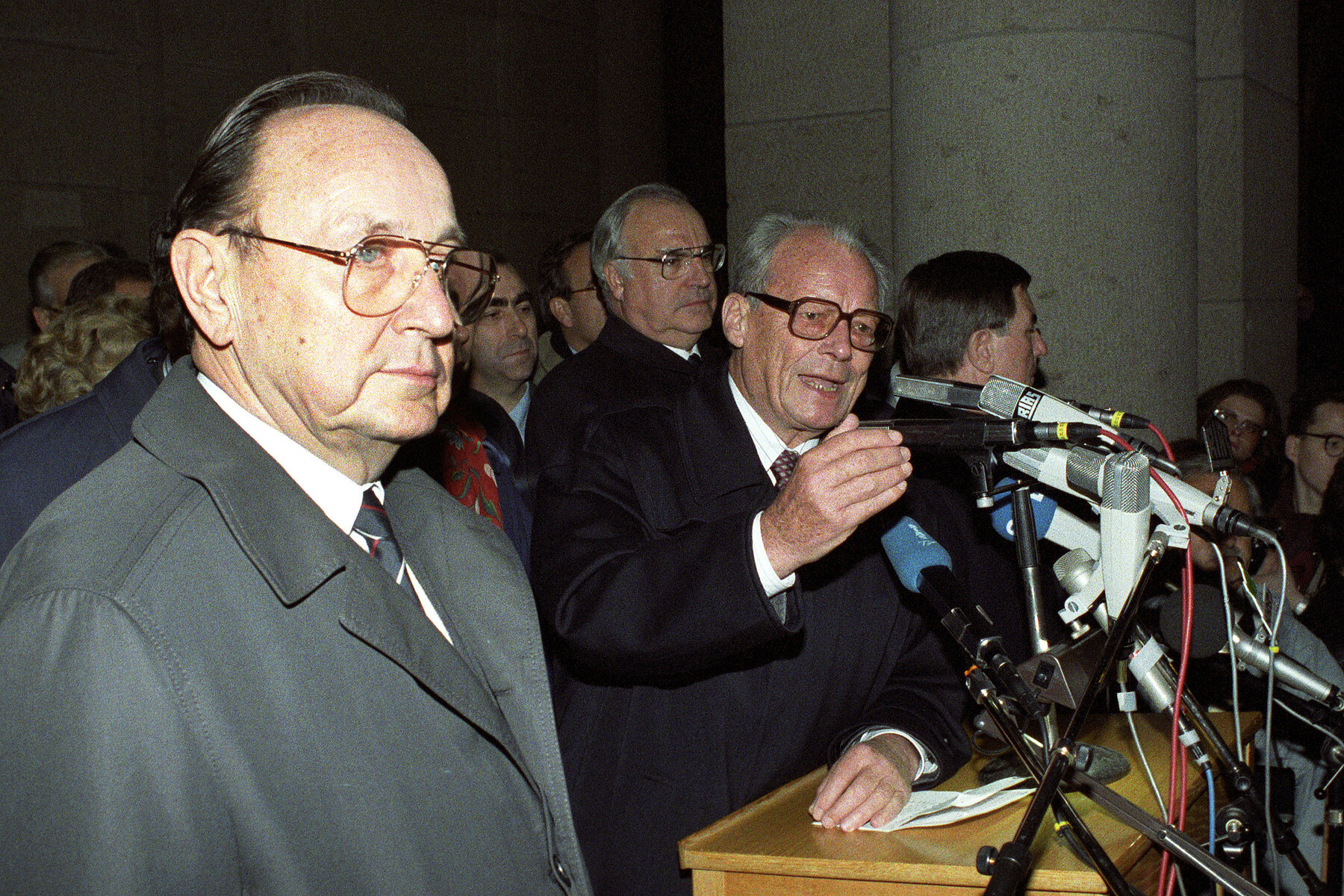 In the front is Hans-Dietrich Genscher, behind him to the right is Helmut Kohl with other politicians. Next to him on the right Willy Brandt speaks into a microphone on a lectern. 