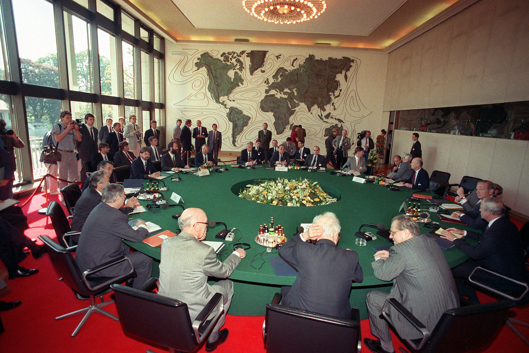 Several men in suits sit around a large green round table. On the left side are journalists taking photographs. 