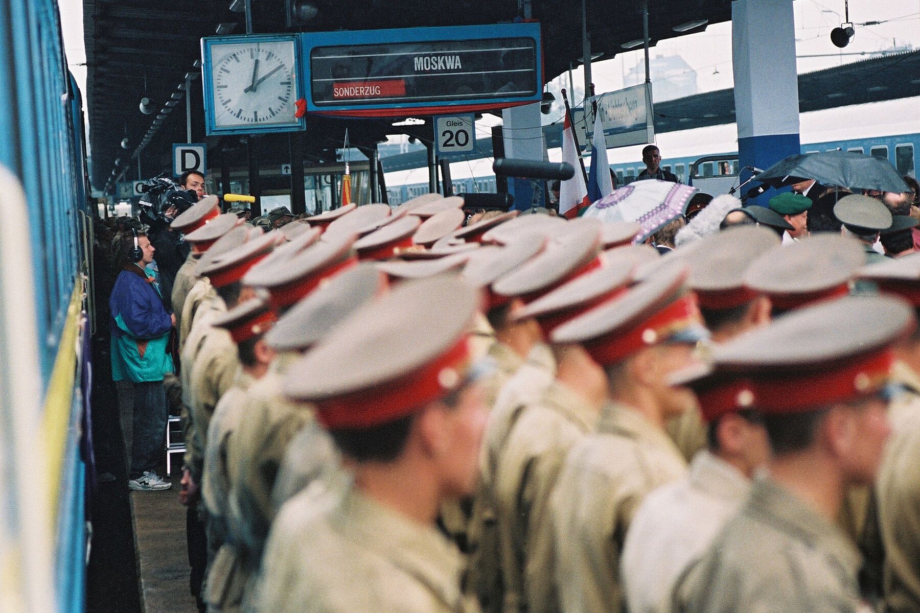 Soldiers in khaki uniform shirts with black and red caps stand on a platform. Above them is a display board with a clock.