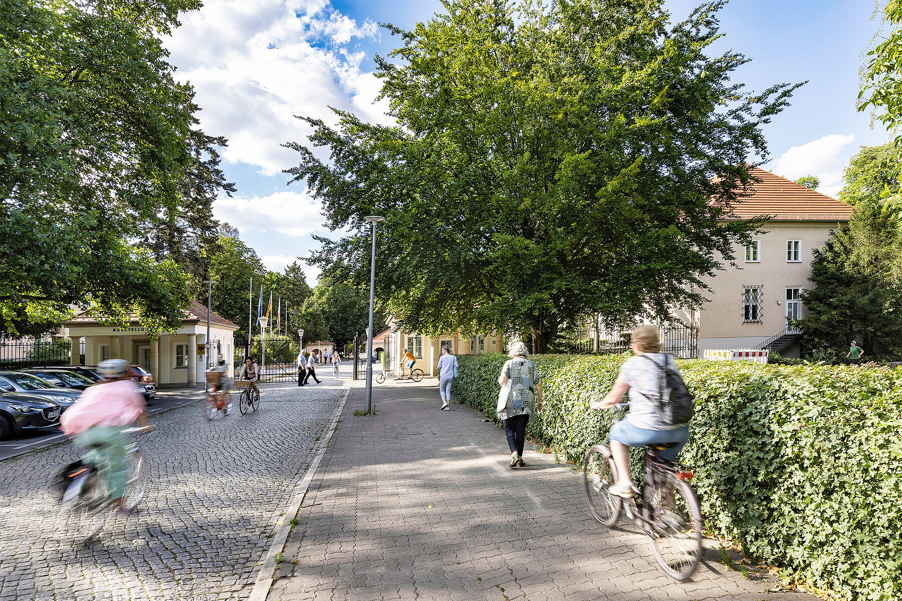 Straße, die auf den Eingang zum Schlosspark zuläuft. Passierende sind zu Fuß und auf dem Fahrrad unterwegs.