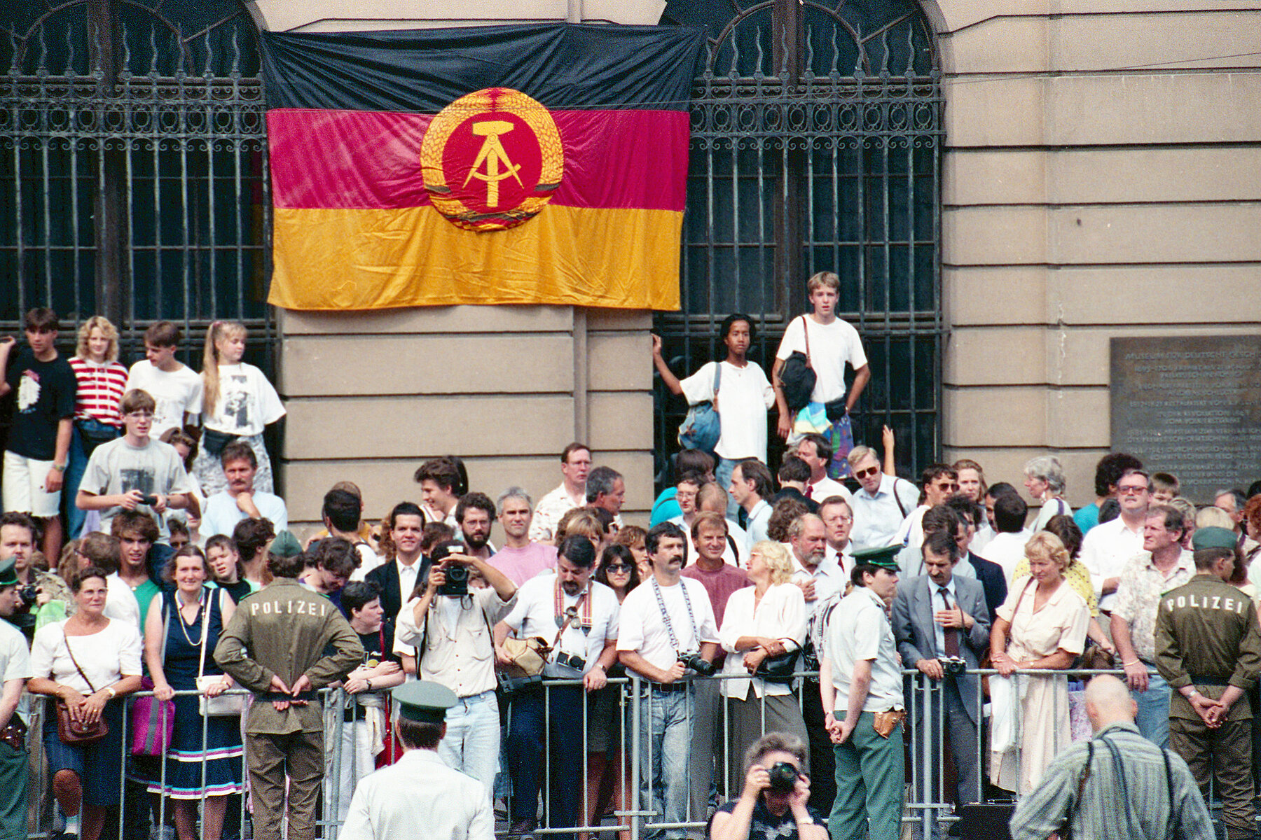 Mehrere Menschen in Freizeitkleidung stehen hinter einer Absperrung. Davor befinden sich Polizisten in Uniform. An einer Wand im Hintergrund hängt die Staatsflagge der DDR.