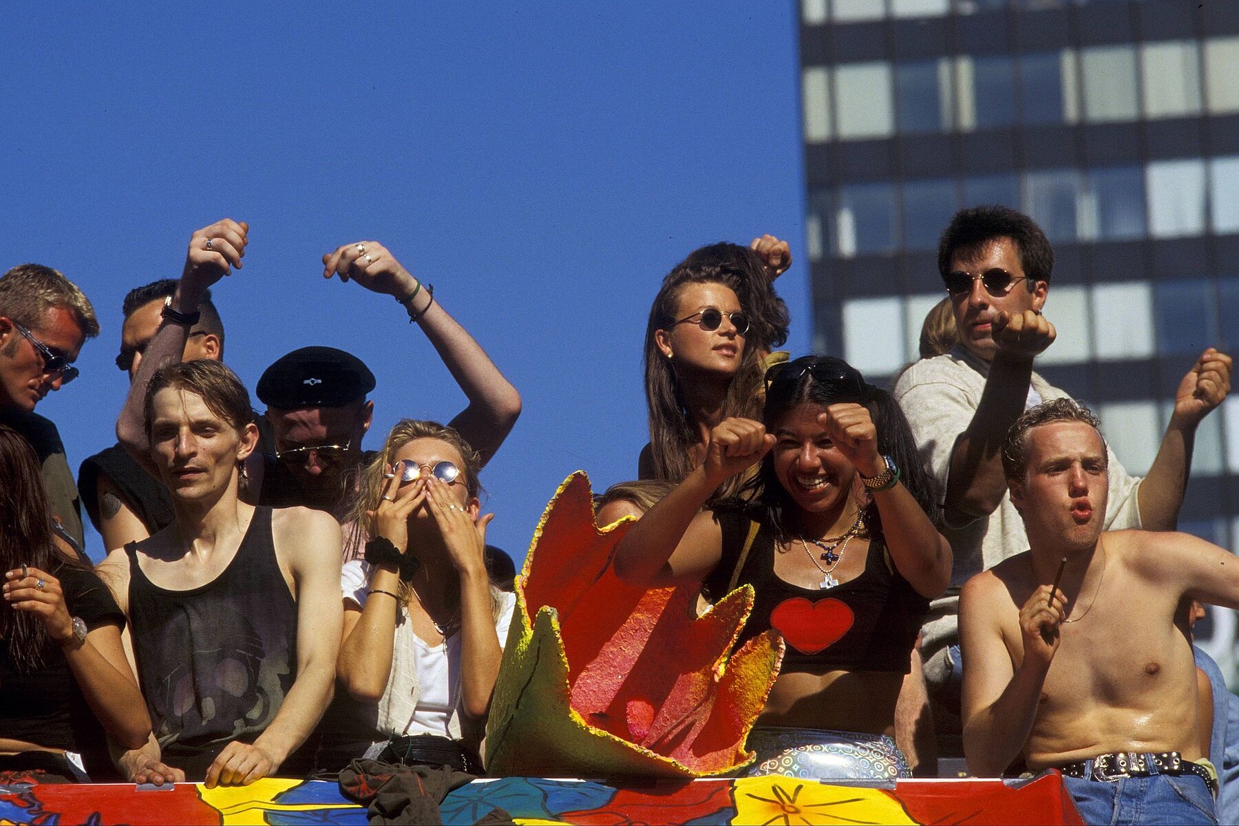 Nine young people celebrate on a wagon under blue sky. In the background on the right is a building.