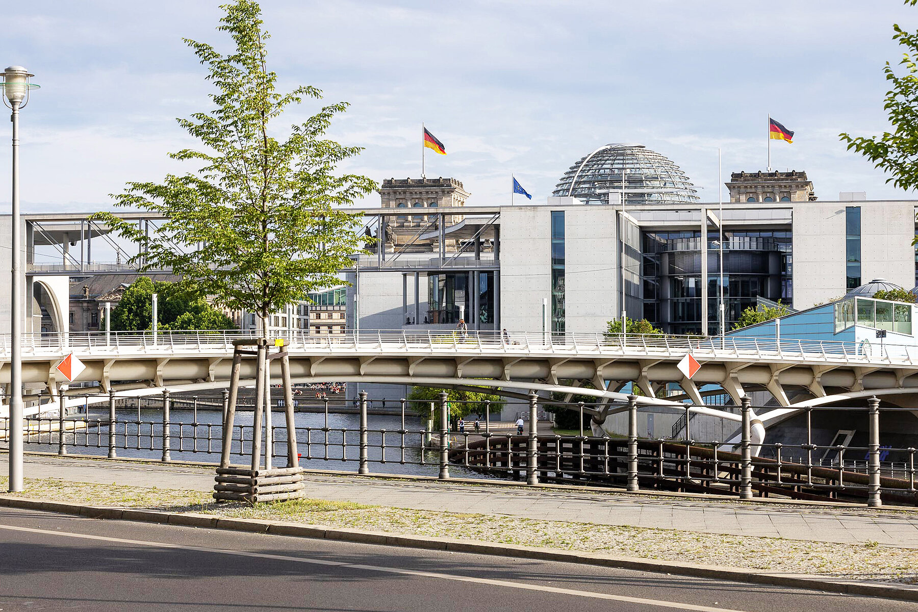 The roof with the dome of the Reichstag protrudes behind a modern government building. In front is the Spree, which is spanned by a bridge.