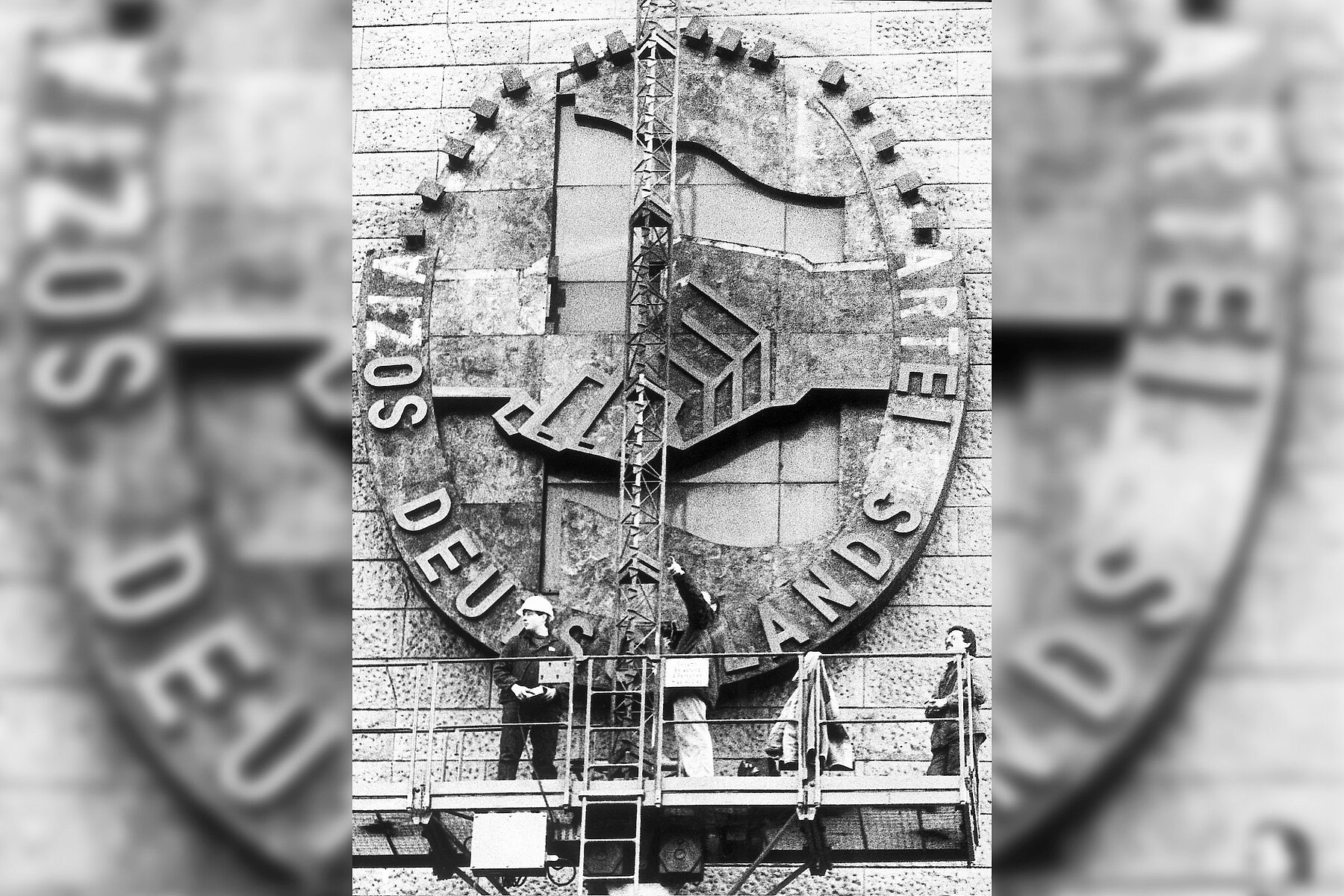 Three workers on a scaffold remove a symbol that is made of stone and several metres high from the facade of the Haus am Werderschen Markt. The symbol depicts the words Socialist Party of Germany and a handshake. 