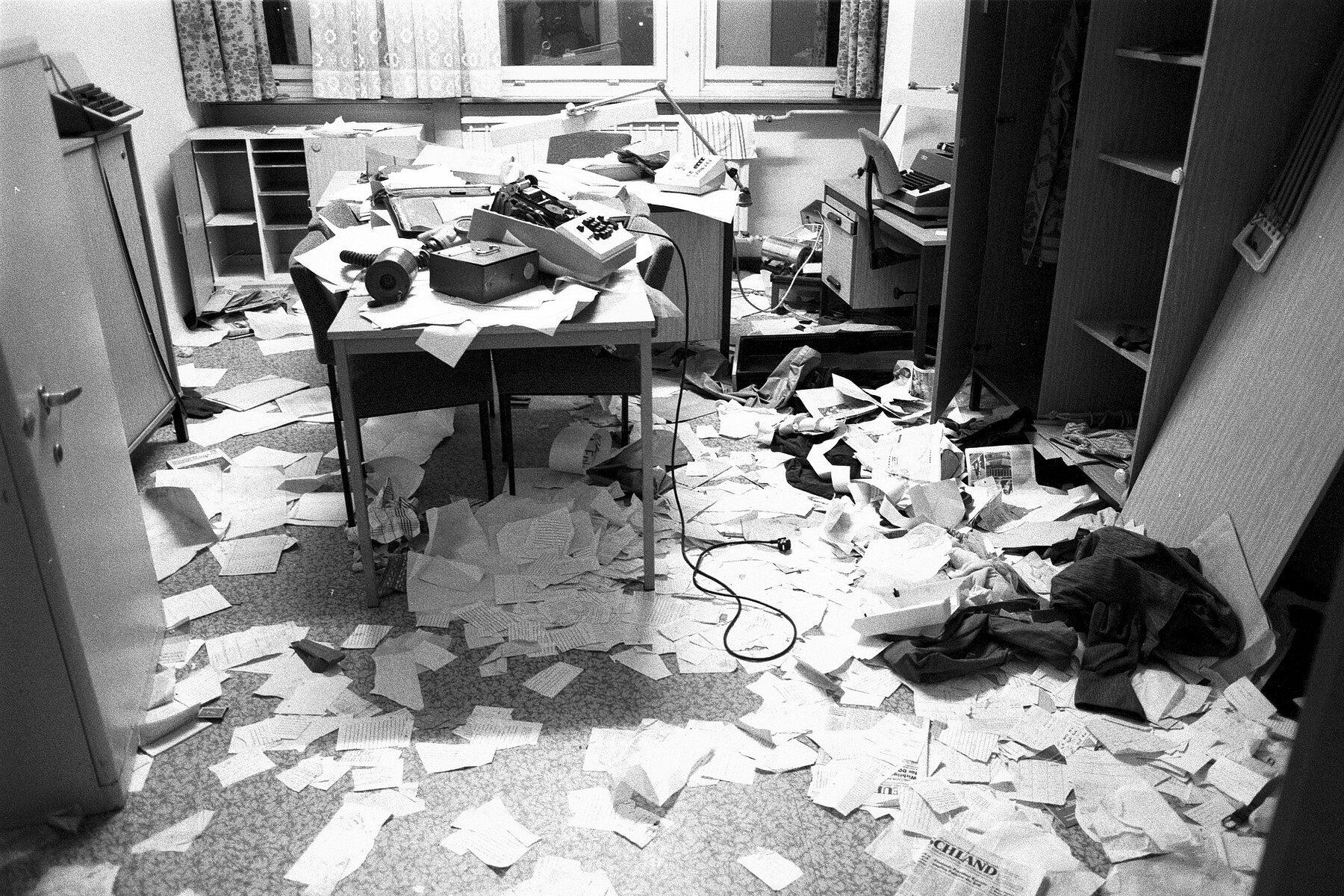 A trashed office with paper on the floor, open cabinets and various writing devices on tables. 