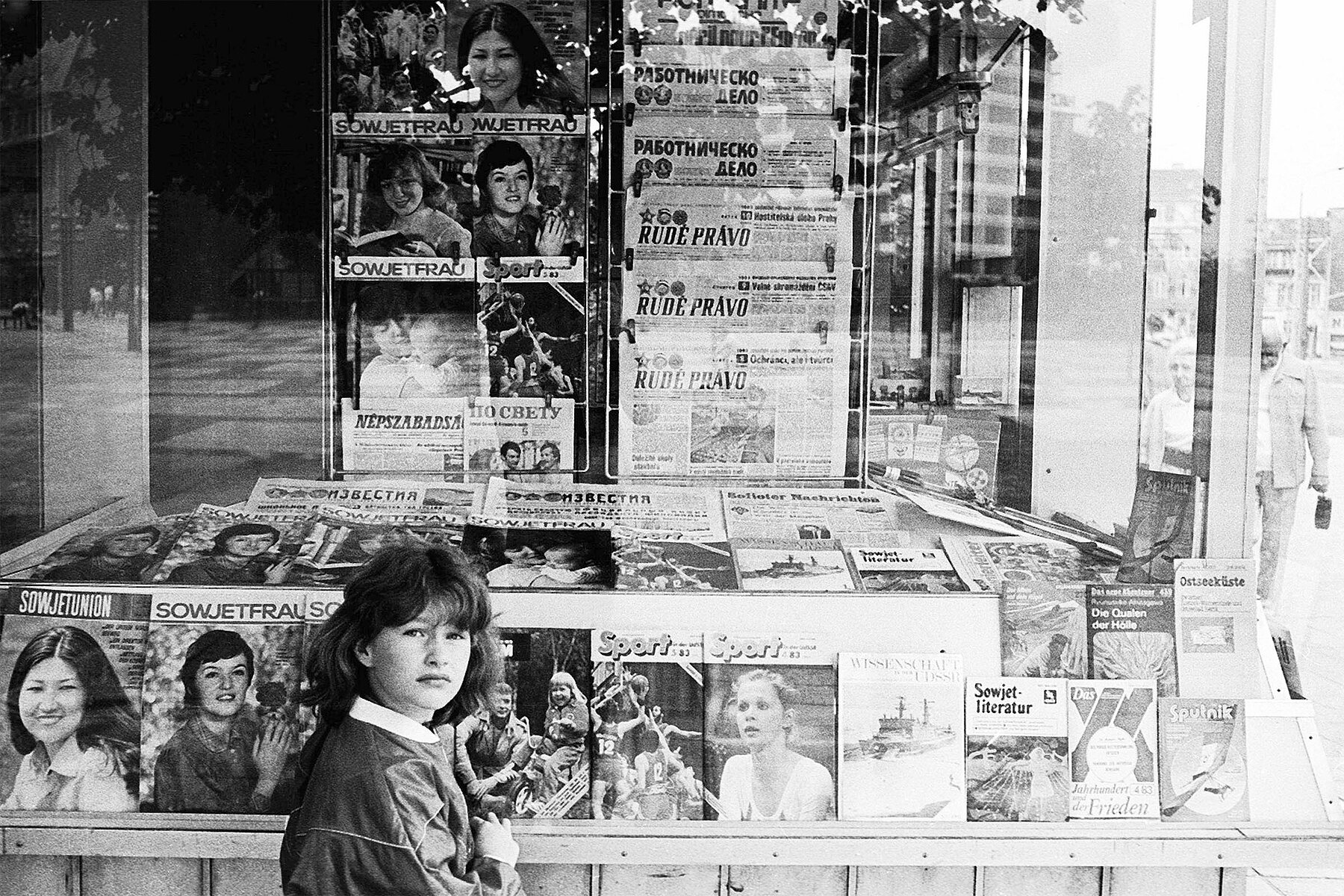 Eine junge Frau steht vor einem DDR-Zeitungskiosk. In der Auslage liegen diverse sozialistische Zeitschriften und Zeitungen.