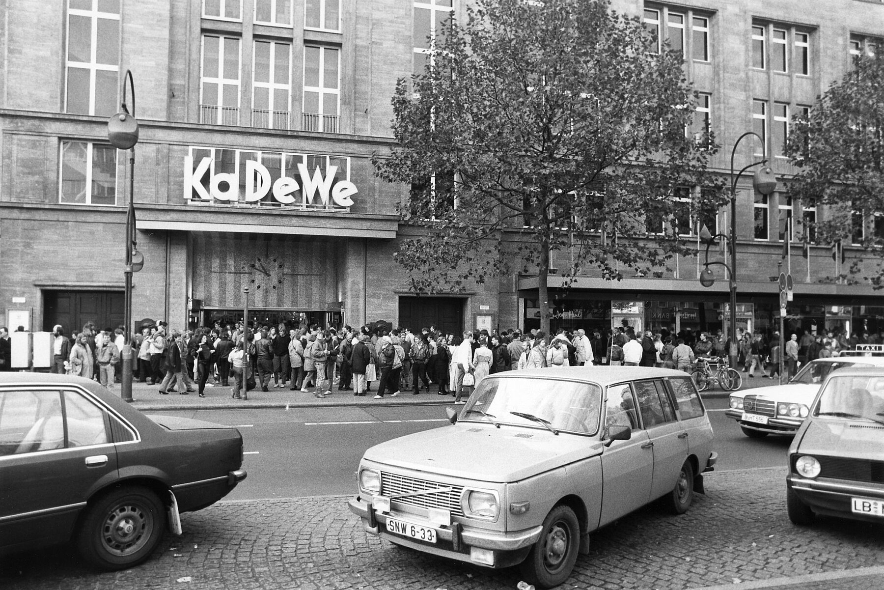 A long line of people in front of an entrance, above it the lettering KaDeWe. In the foreground is a street and parked cars. 