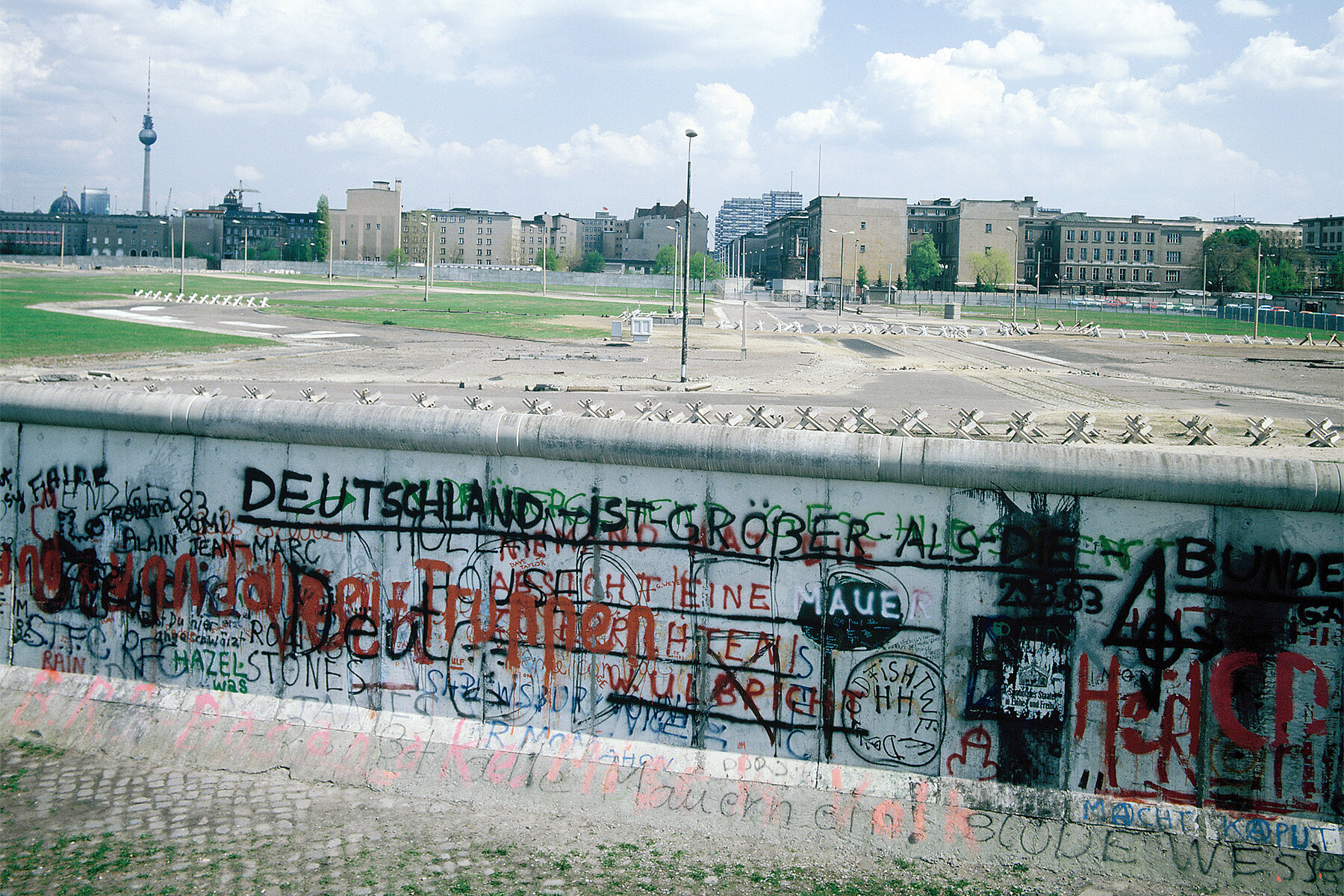 Die Mauer am Potsdamer Platz. Sie ist mit Schriftzügen in schwarz und rot bemalt dahinter liegt eine Brachfläche.