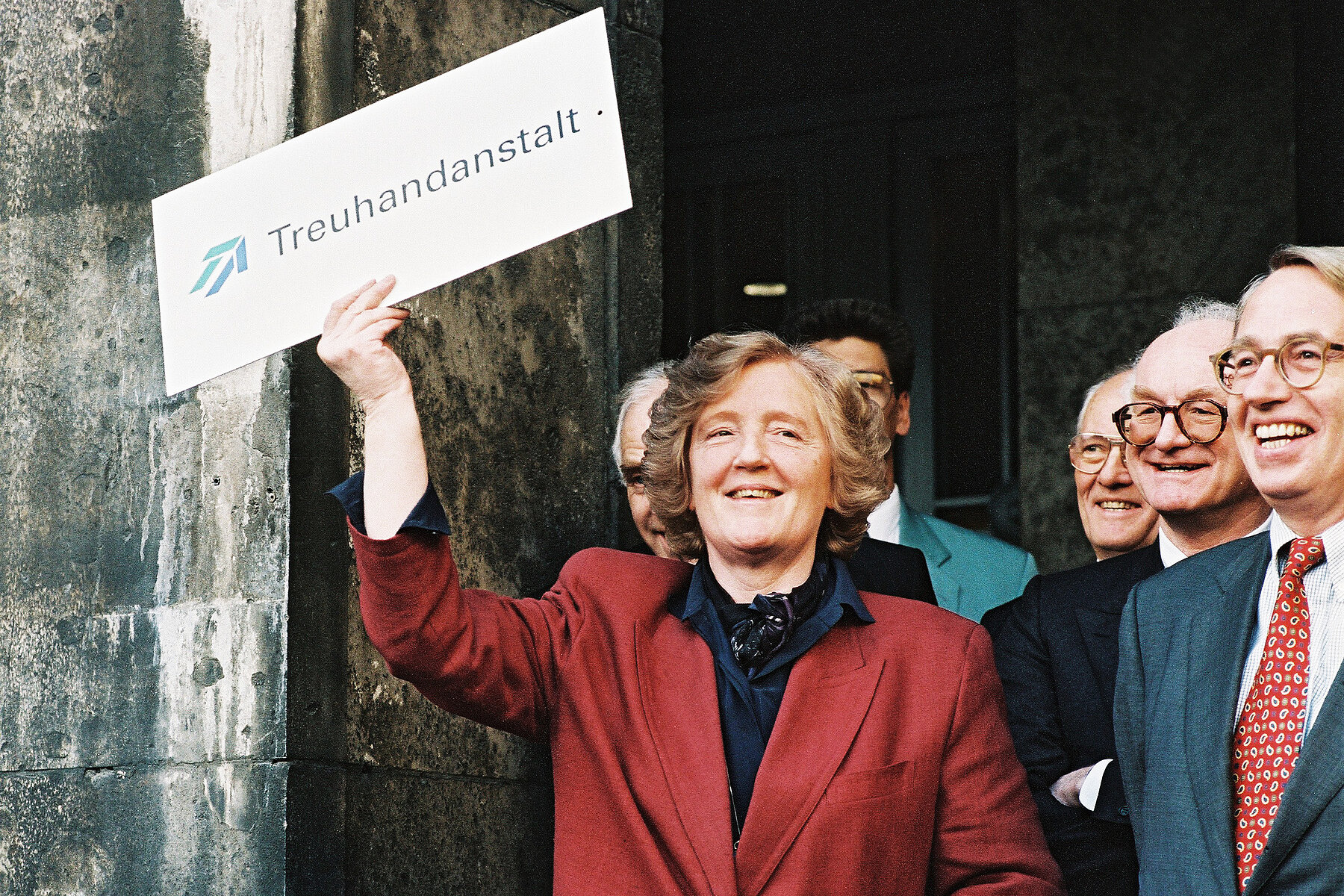 Treuhand President Birgit Breuel wears a red blazer. In her right hand she holds a sign saying Treuhand. Men in suits stand to her right.