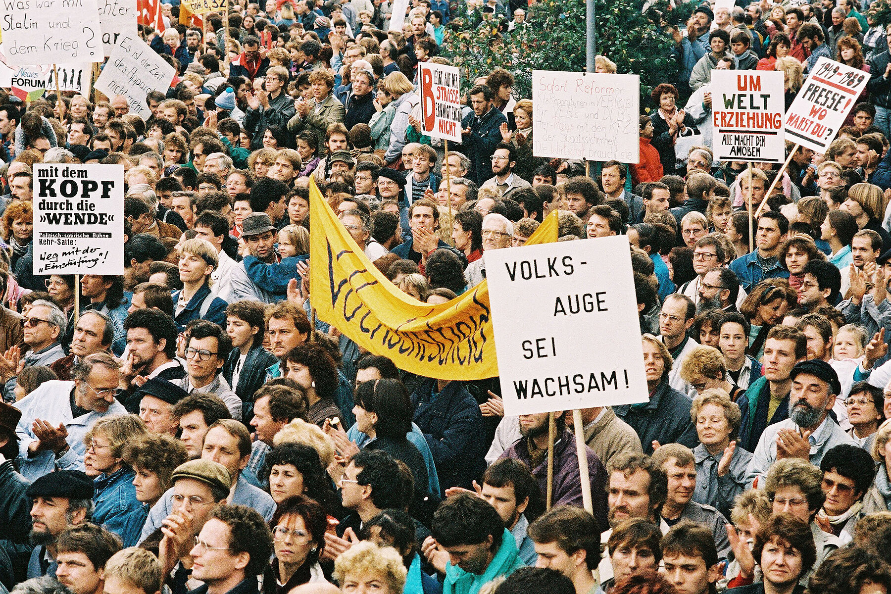 A large crowd of demonstrators on Alexanderplatz, some carry signs. A sign in the foreground reads People's eye, be vigilant.