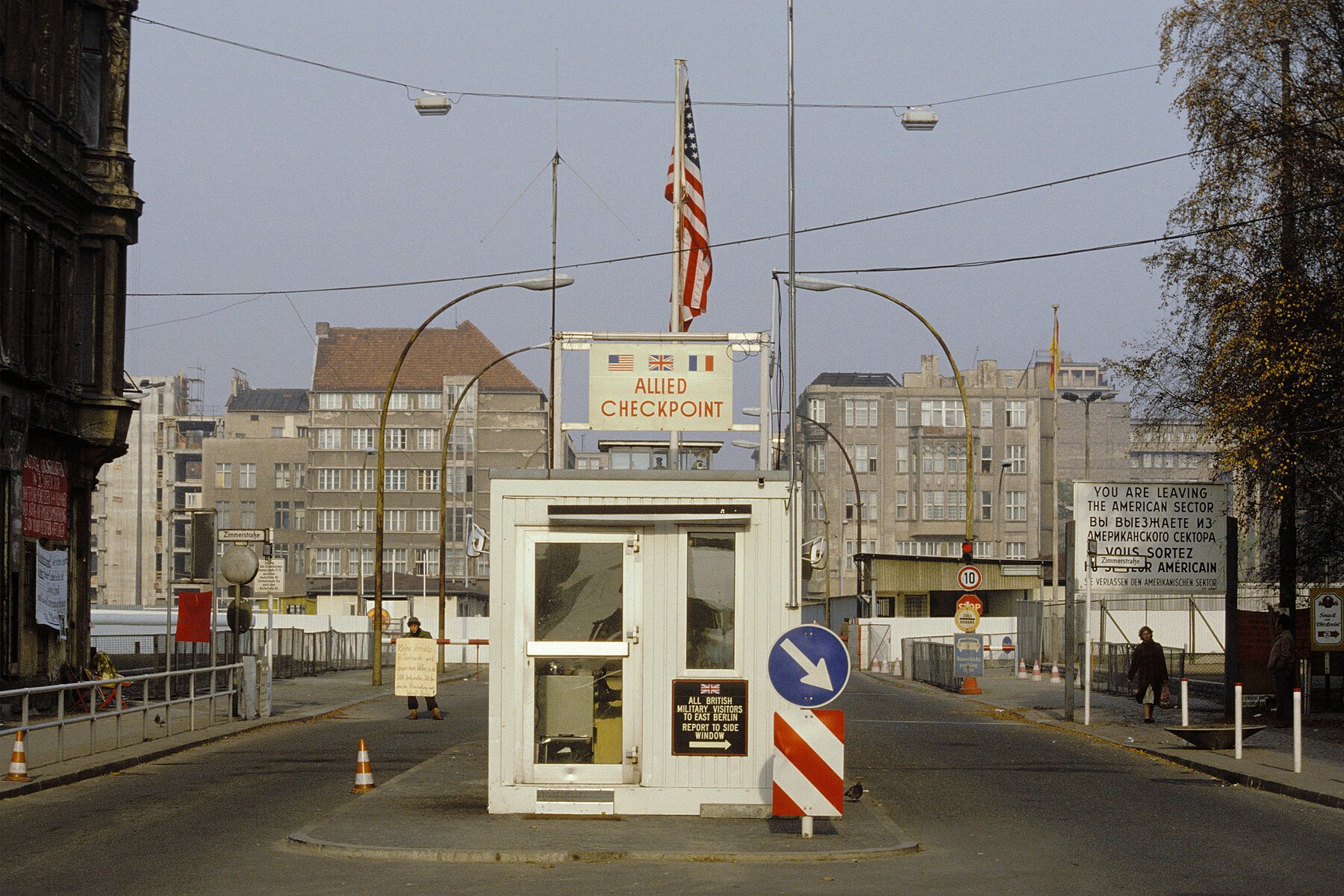 On the control booth there is a sign with the inscription: Allied Checkpoint. Above it hangs an American flag. On the right side of the booth, a sign reads: All British Military Visitors To East Berlin Report To Side Window. In the background on the right a sign reads in English, Russian, French and German: You are leaving the American Sector.