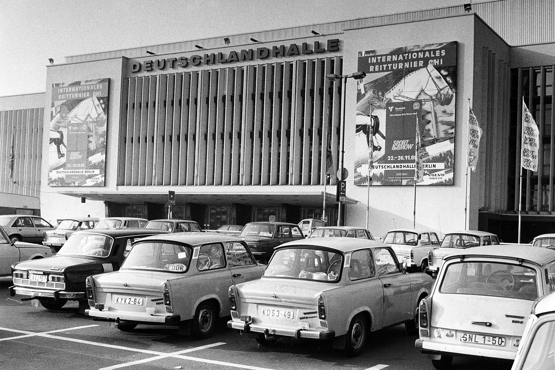 Eine Reihe von Trabants parkt vor einem Gebäude mit dem zentralen Schriftzug Deutschlandhalle. Links und rechts des Gebäudes sind Plakate zu sehen, die für ein internationales Reitturnier im November 1989 werben.