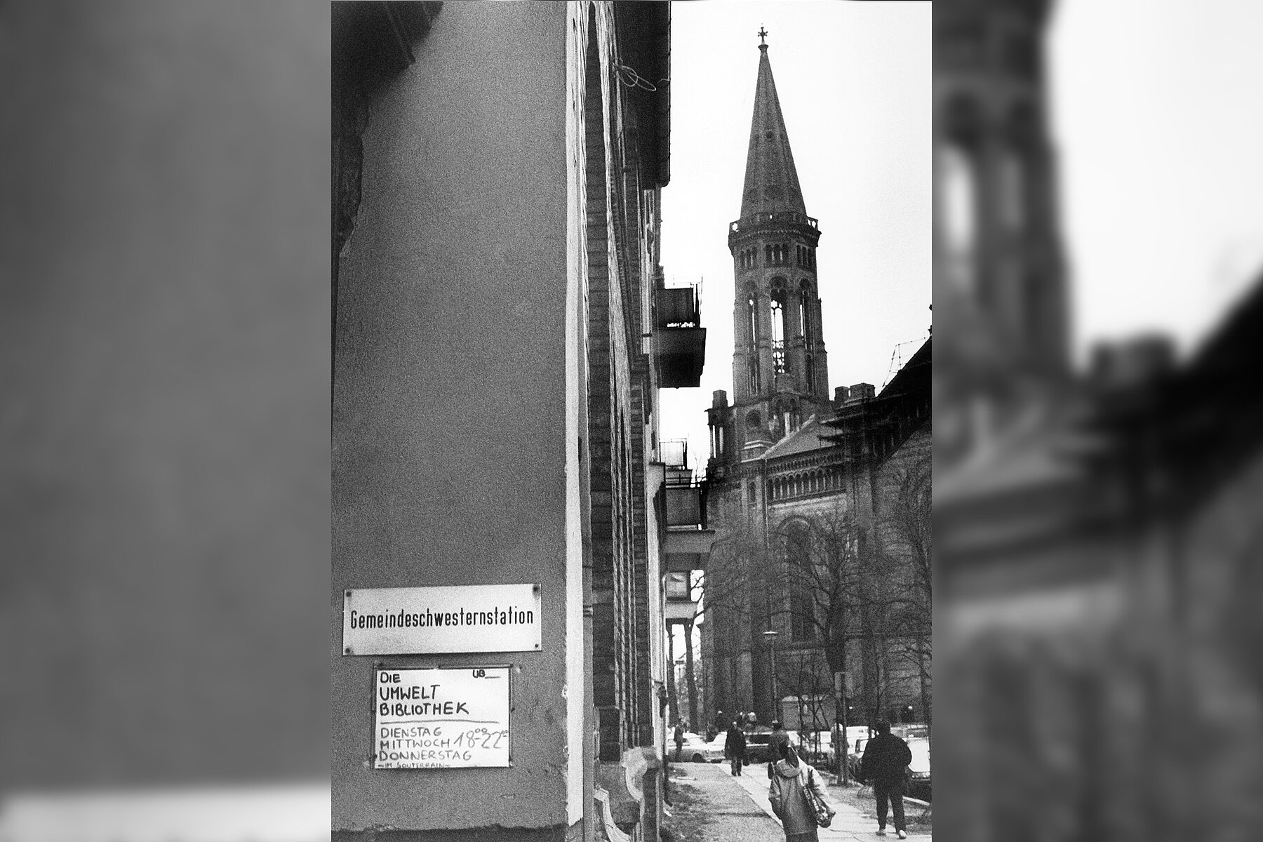 Auf einem Schild im Hauseingang des Gemeindehauses der Zionskirche stehen handschriftlich die Öffnungszeiten der Umweltbibliothek, rechts vom Eingang befindet sich die Zionskirche.