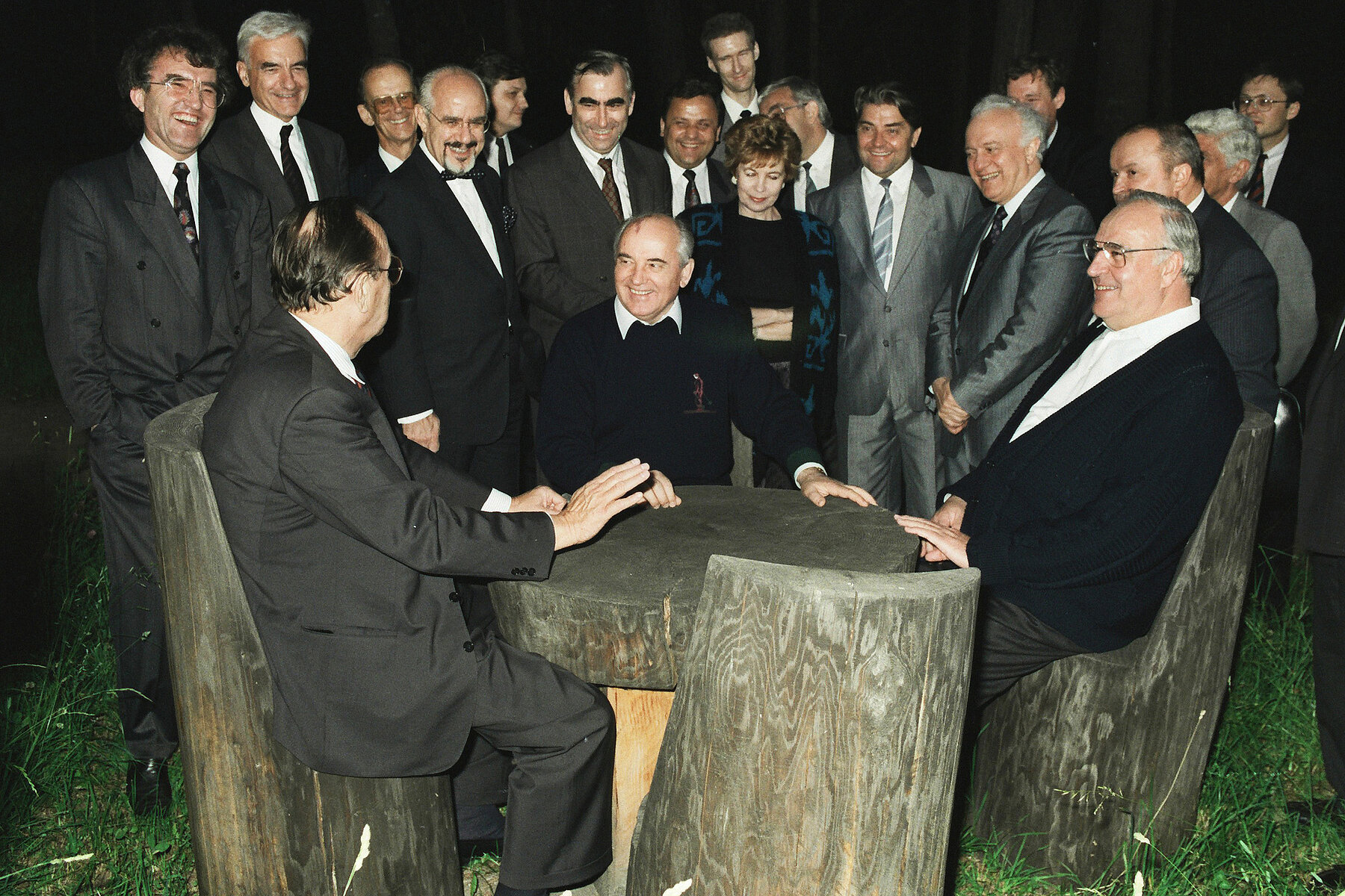 Three men are seated at a round wooden table. Behind them are several men and a woman in suits. It's night.