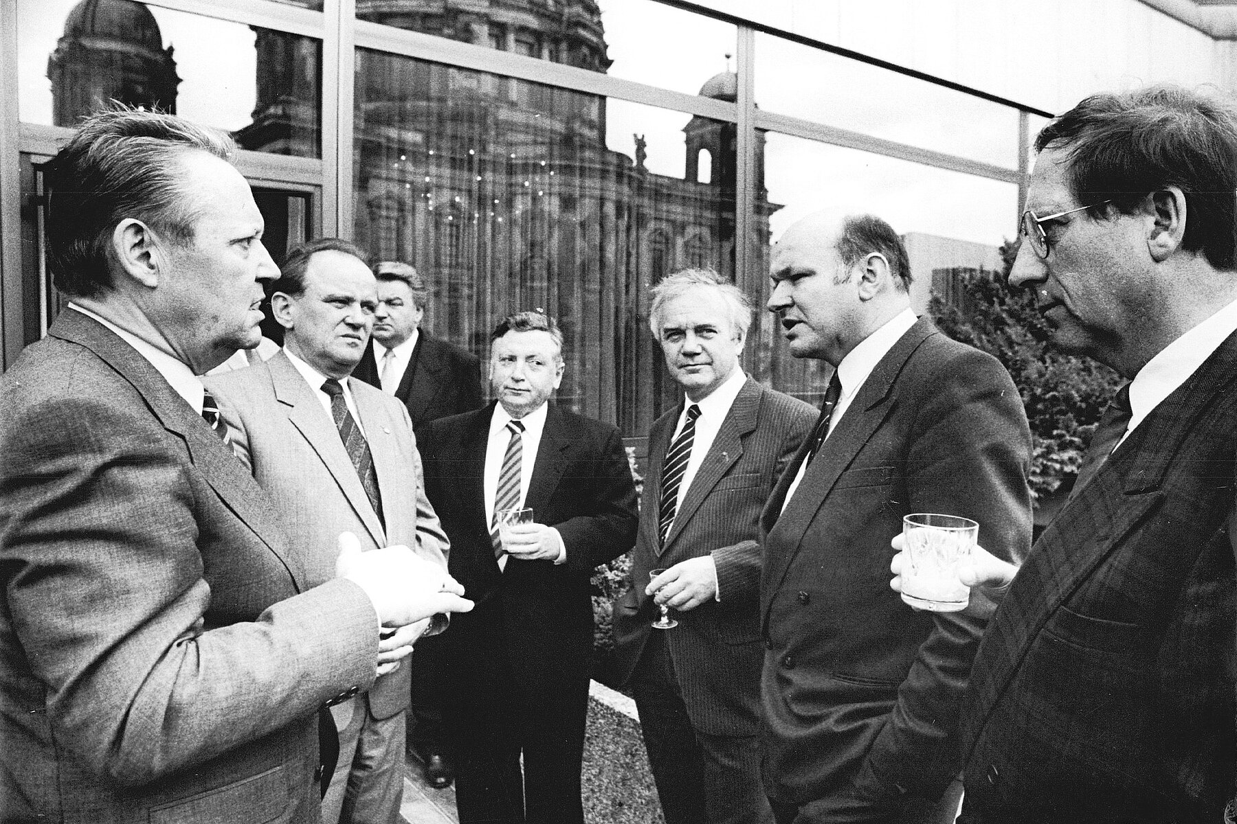 Sieben Männer in Anzügen im Gespräch vor einer Fensterfront in der sich der Berliner Dom spiegelt.