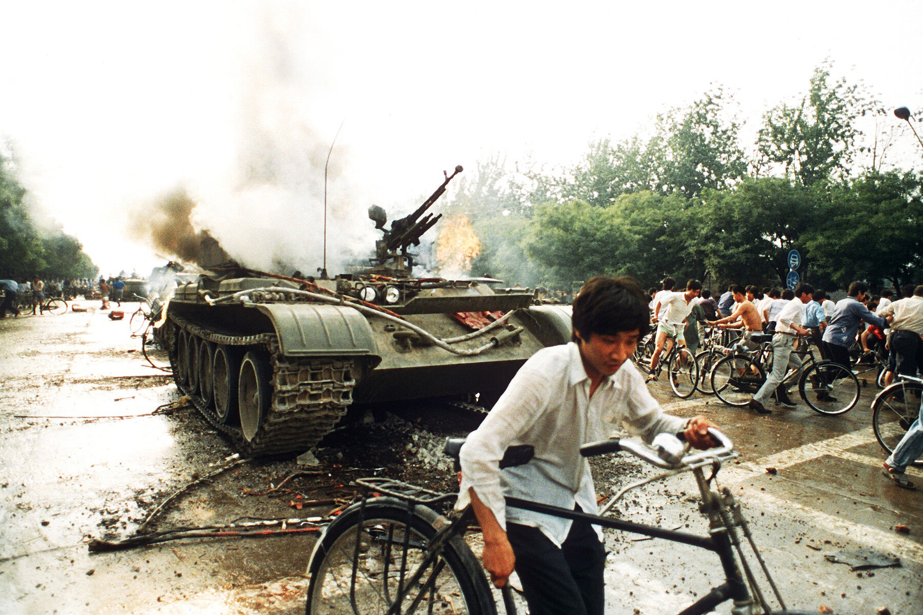 A smoking tank on a street, with a man pushing his bike in the foreground.