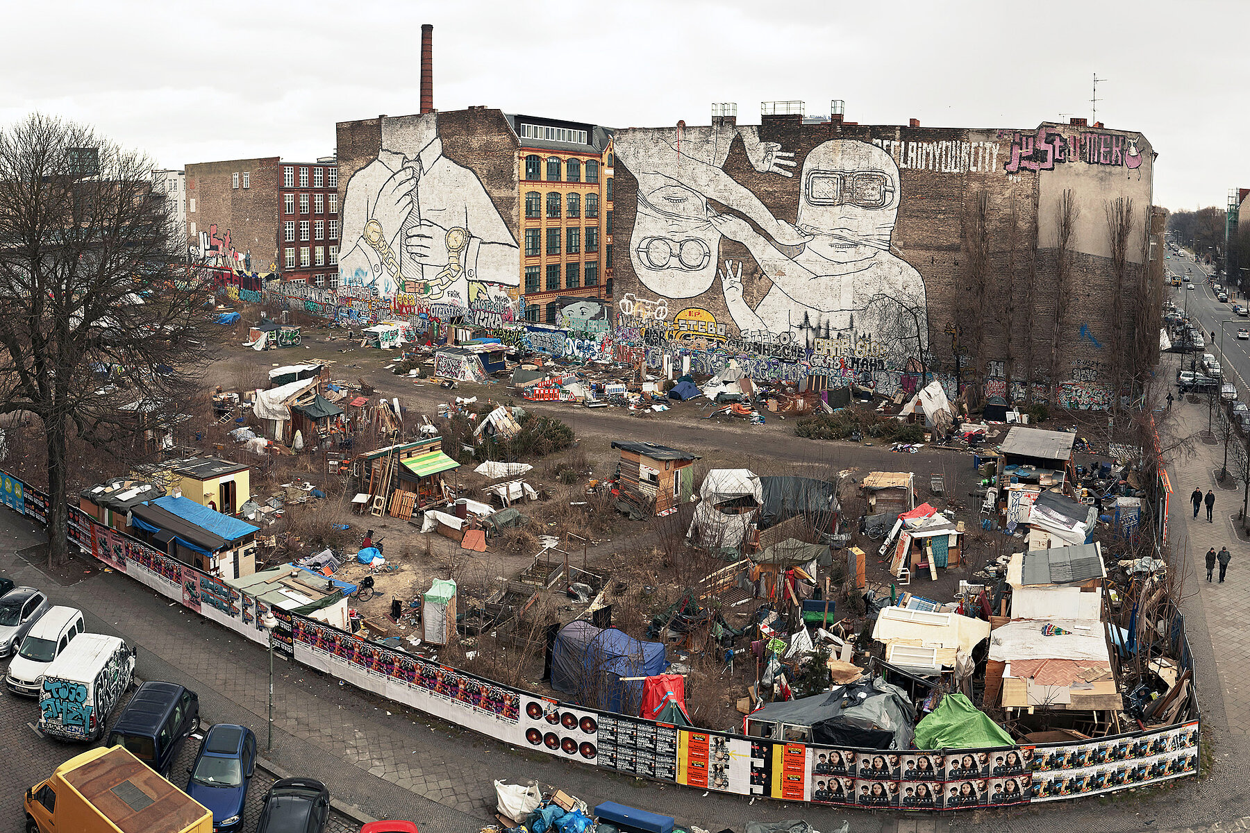 The wasteland on Cuvrystraße from above. A tent city stands on the open space, and on each of the two house walls in the background is a mural with white figures.