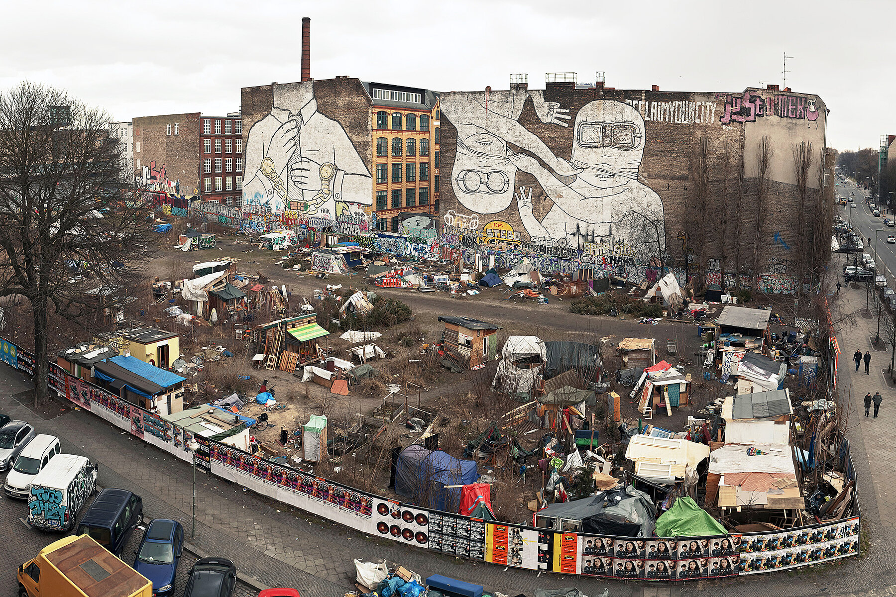 Die Brachfläche an der Cuvrystraße von oben. Auf der Freifläche steht eine Zeltstadt, auf den beiden Hauswänden im Hintergrund befindet sich jeweils ein Wandbild mit weißen Figuren.