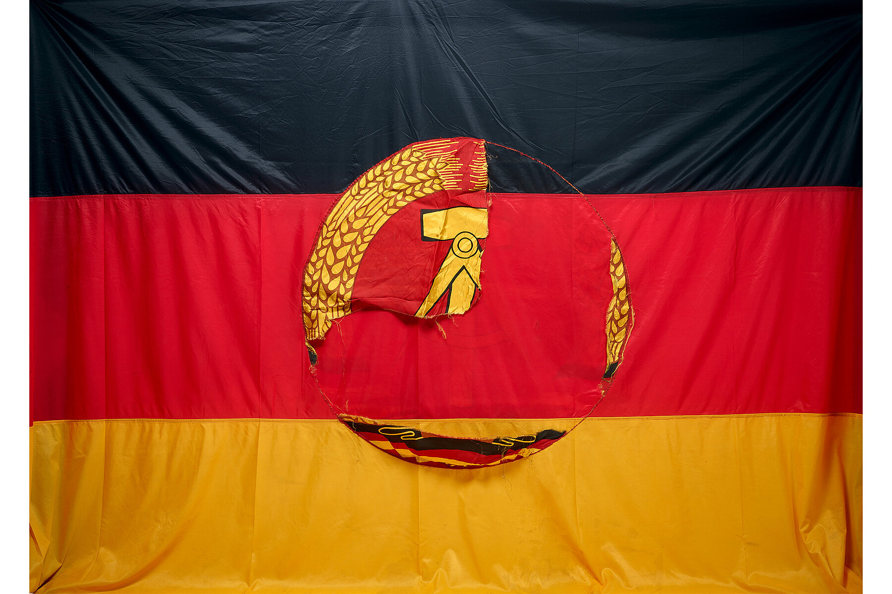 A black-red-gold striped state flag of the GDR with the symbol of the hammer and sickle in the centre, partly torn off.
