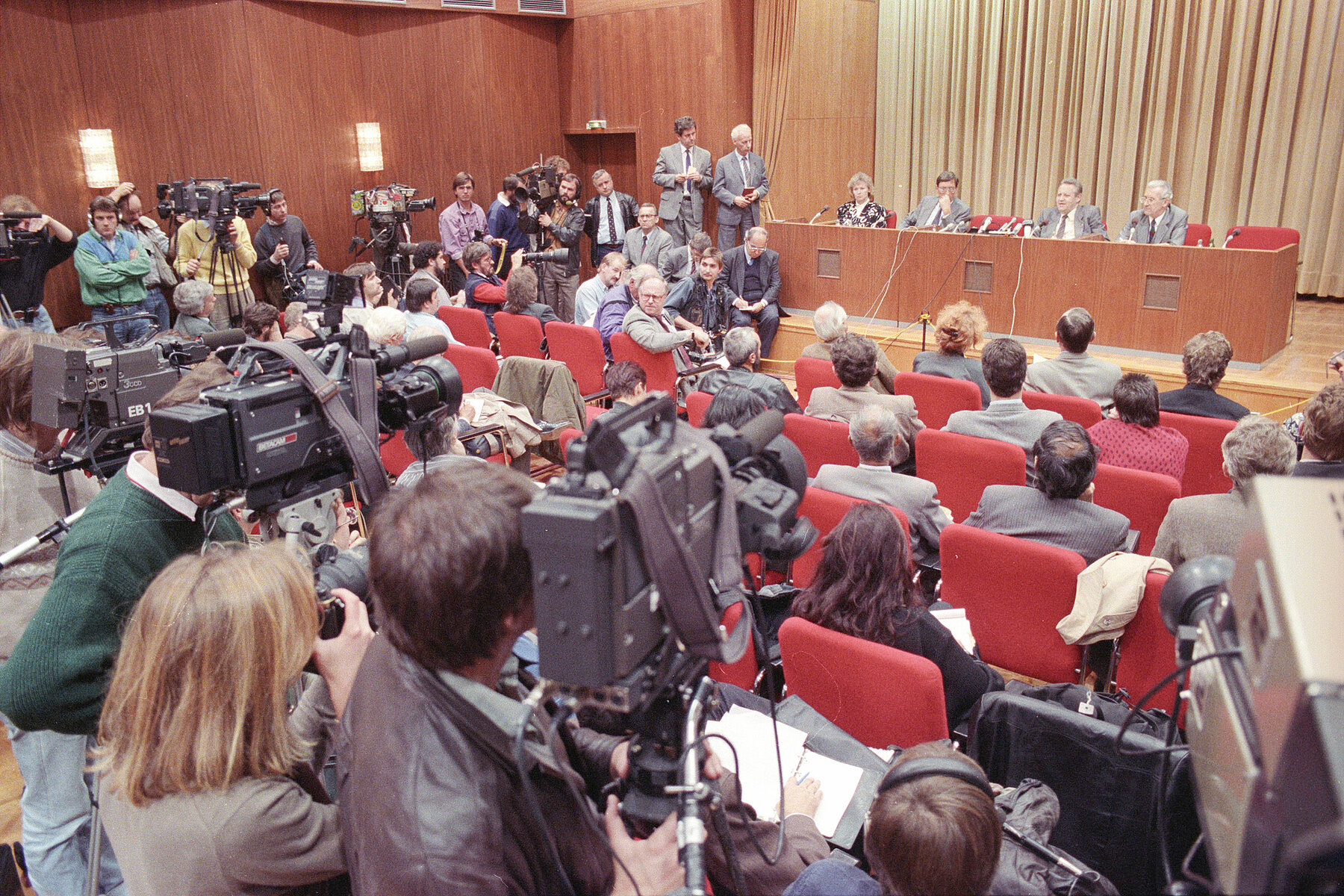 Three men and one woman sit behind a desk with microphones. Journalists sit and stand in front of them with film and photo cameras. 