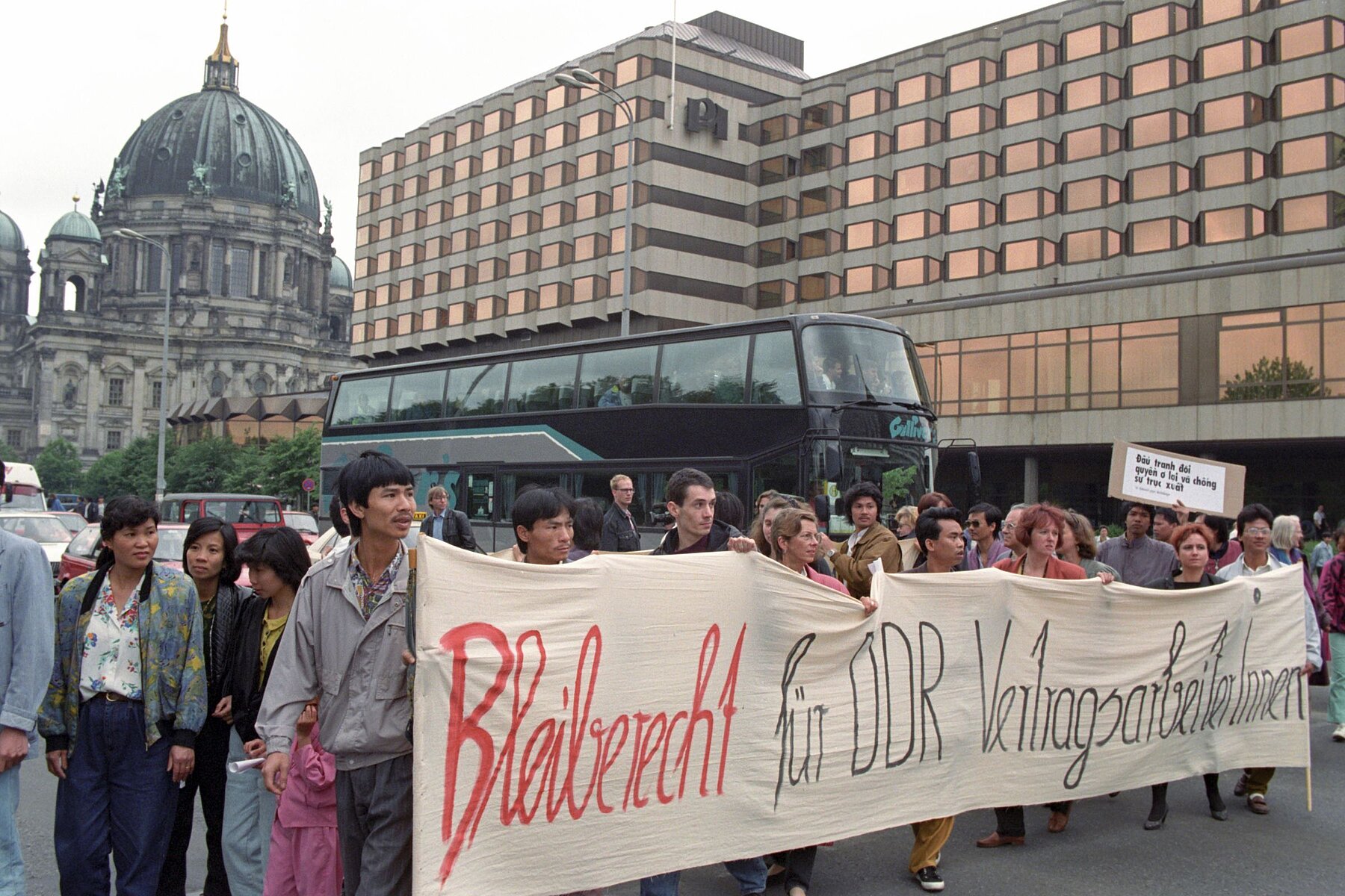 A group of people holds a banner that reads Bleiberecht für DDR VertragsarbeiterInnen, in  English Right to stay for GDR contract workers. To the right of them is a double-decker bus, behind it the Palasthotel, in English Palace Hotel. The Berlin Cathedral looms in the background of the picture. 