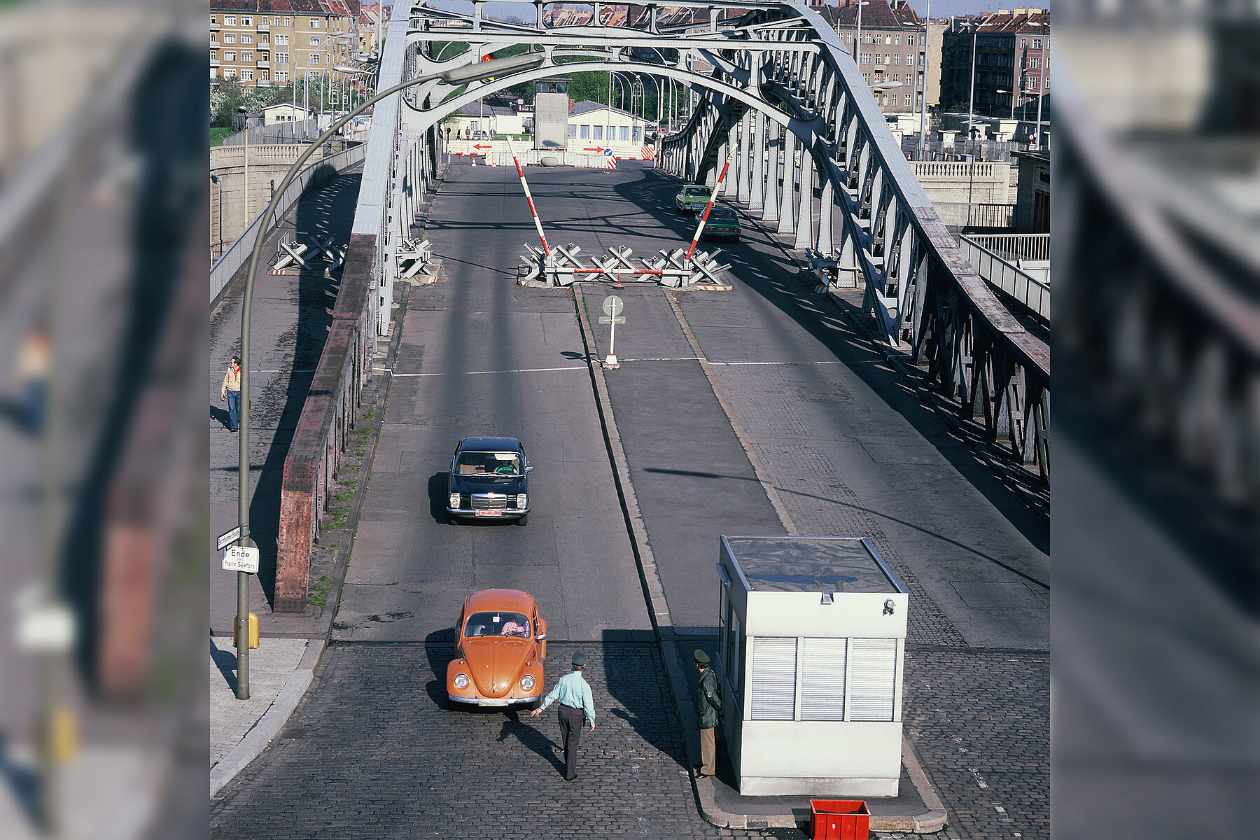 Two cars drive over the Bösebrücke. In the background on the bridge is a border barrier. 