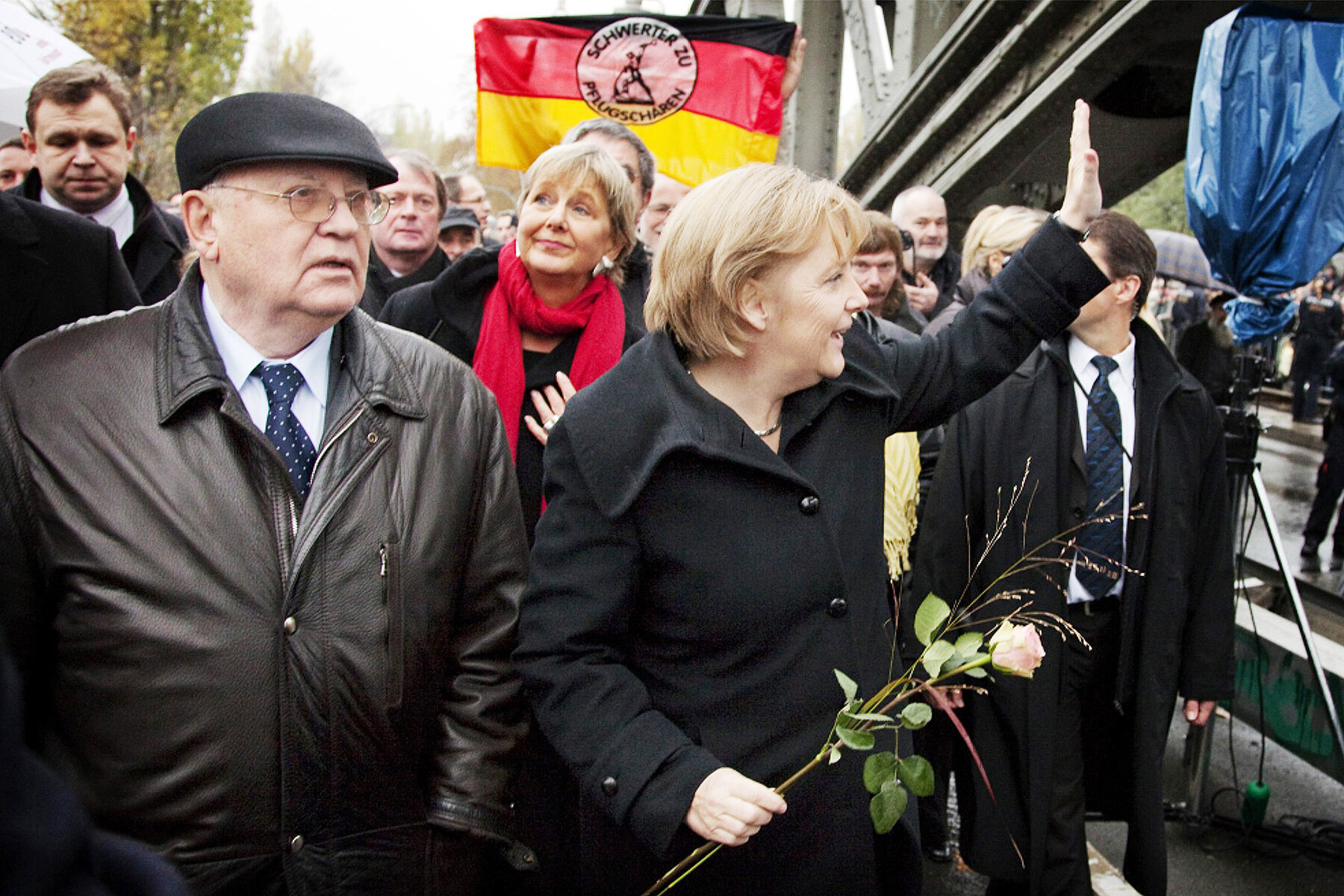 Michael Gorbatschow links und Angela Merkel rechts an der Bernauer Straße auf der Bösebrücke. Merkel hält eine Rose in der Hand. Hinter ihnen wird eine Deutschlandfahne geschwenkt.
